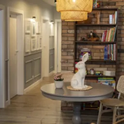 Wnętrze gabinetu: okrągły stół z dekoracjami (królik, kwiaty), regał z książkami na tle ceglanej ściany, widok na korytarz z ramkami na ścianach.