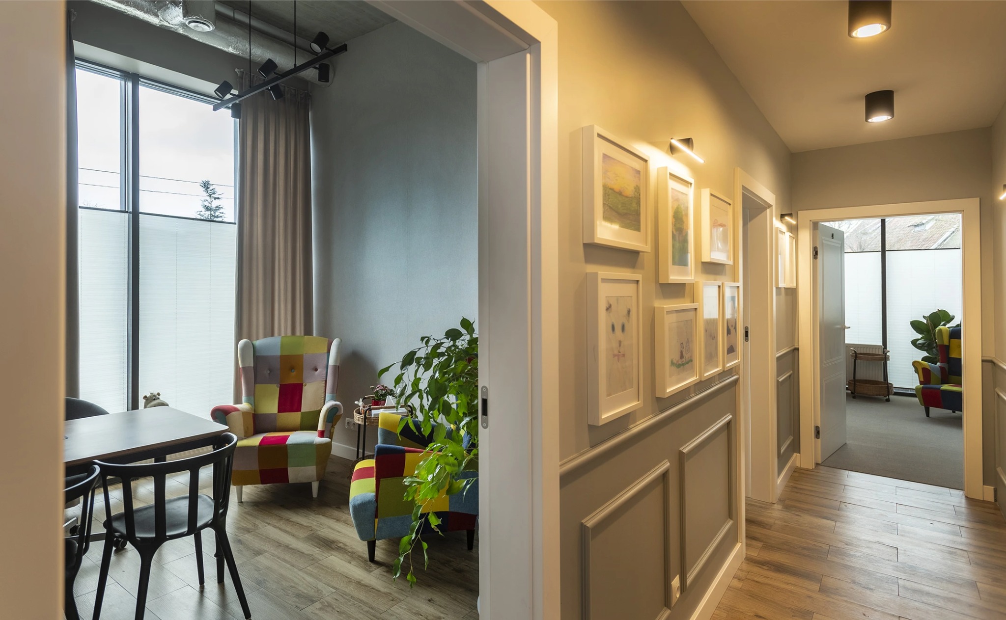Korytarz z galerią obrazów i widokiem na pokój z patchworkowym fotelem. Nowoczesne oświetlenie i drewniana podłoga. Przytulne wnętrze gabinetu.