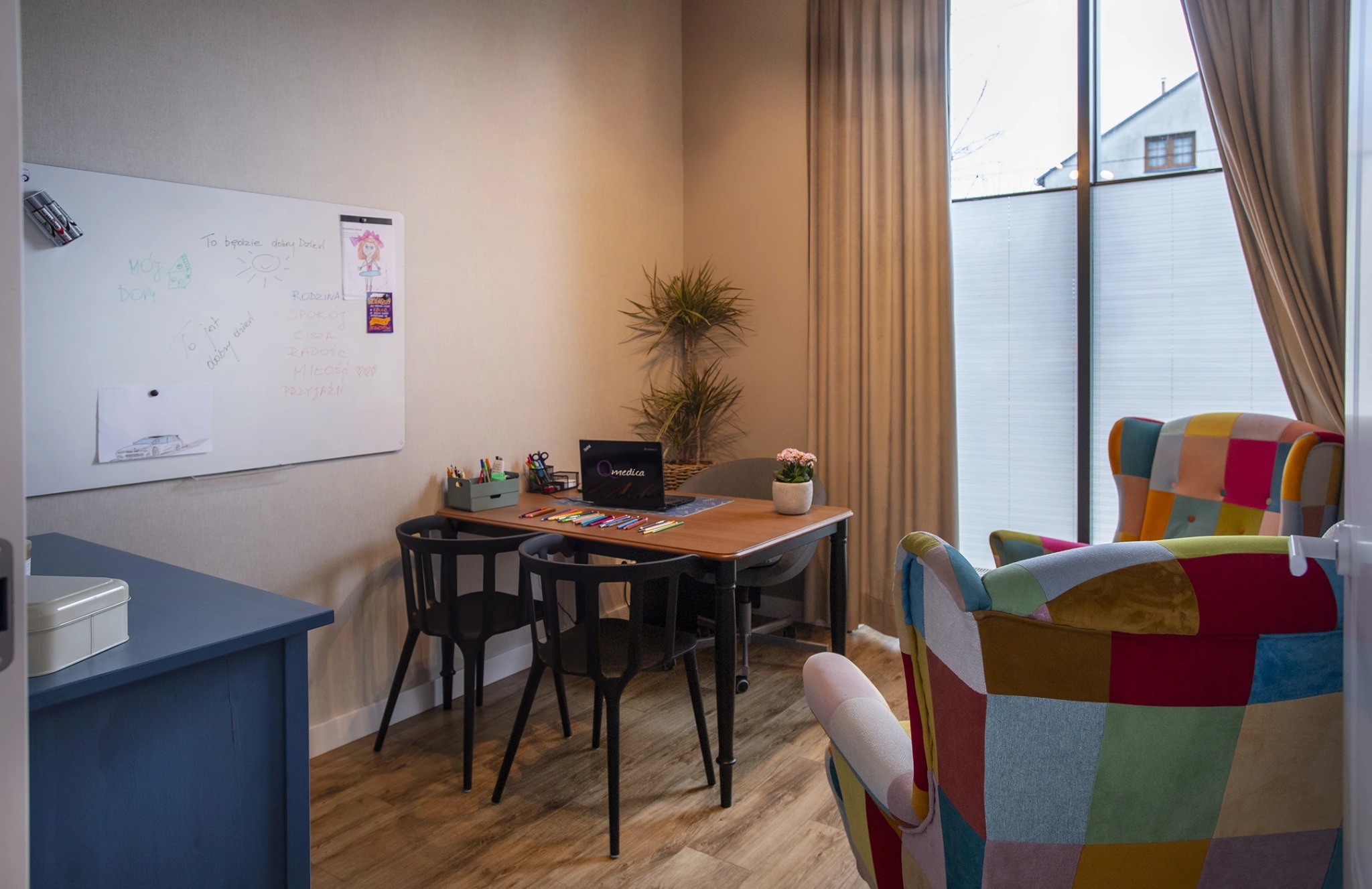 Jasny gabinet z biurkiem, laptopem Qmedica, kolorowymi kredkami, rysunkami na tablicy i patchworkowym fotelem. Okno z żaluzjami w tle.