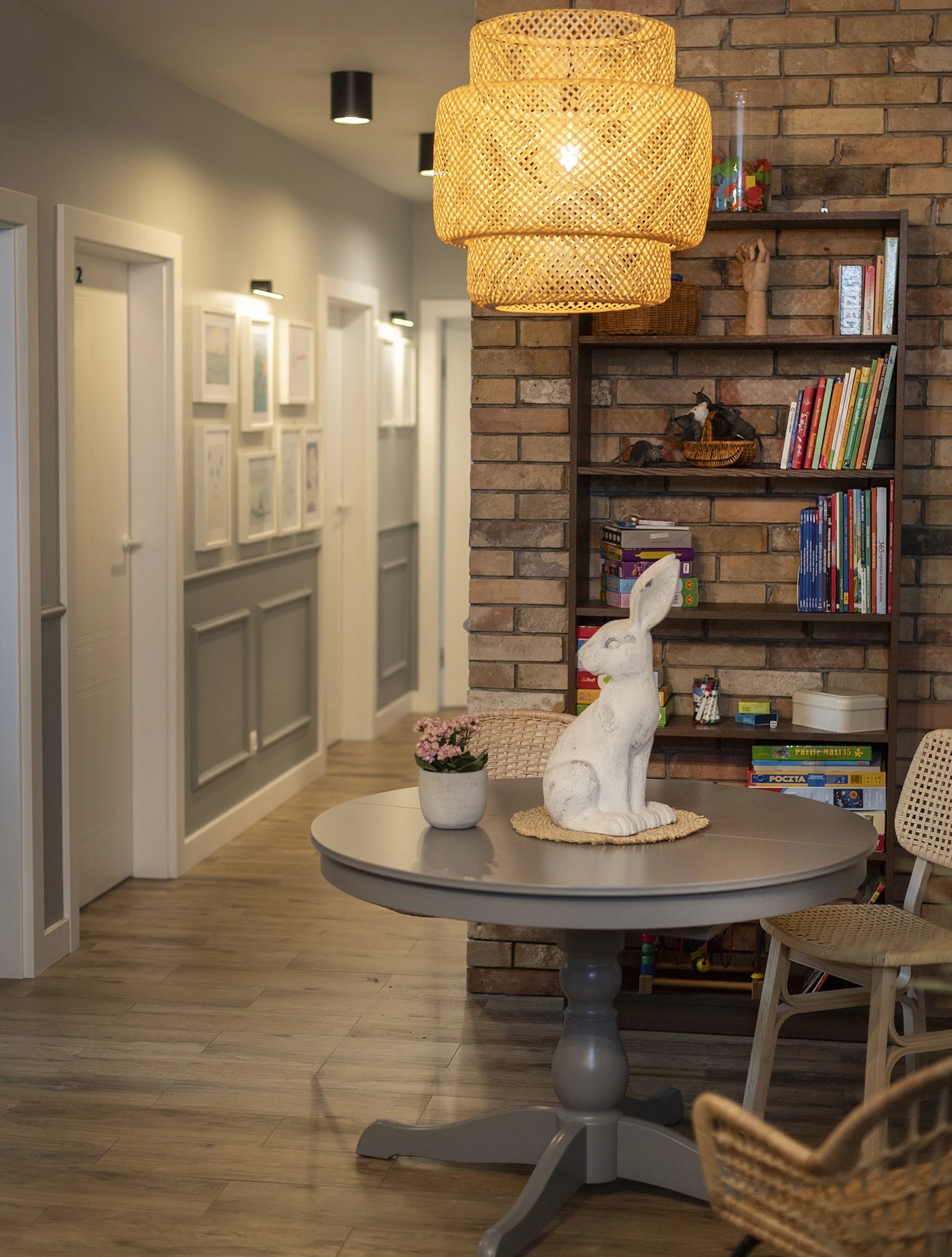 Wnętrze gabinetu: okrągły stół z dekoracjami (królik, kwiaty), regał z książkami na tle ceglanej ściany, widok na korytarz z ramkami na ścianach.