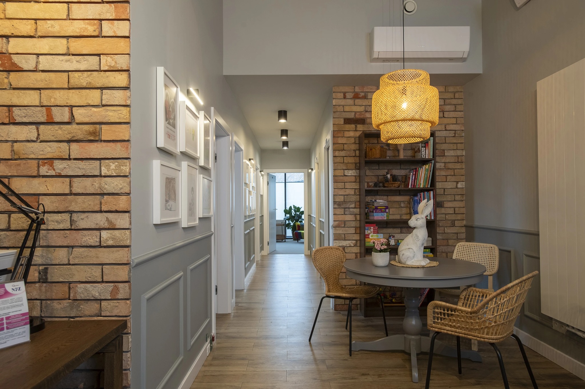 Wnętrze gabinetu: ceglane ściany, drewniana podłoga, okrągły stół z wiklinowymi krzesłami i dekoracyjną lampą, tworzące przytulną atmosferę.