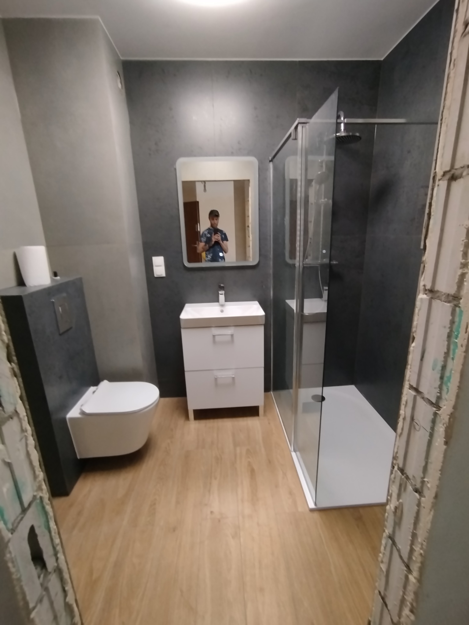 Nowoczesna łazienka po remoncie: szara ściana, biała umywalka z szafką, kabina prysznicowa typu walk-in. W lustrze odbicie osoby robiącej zdjęcie.