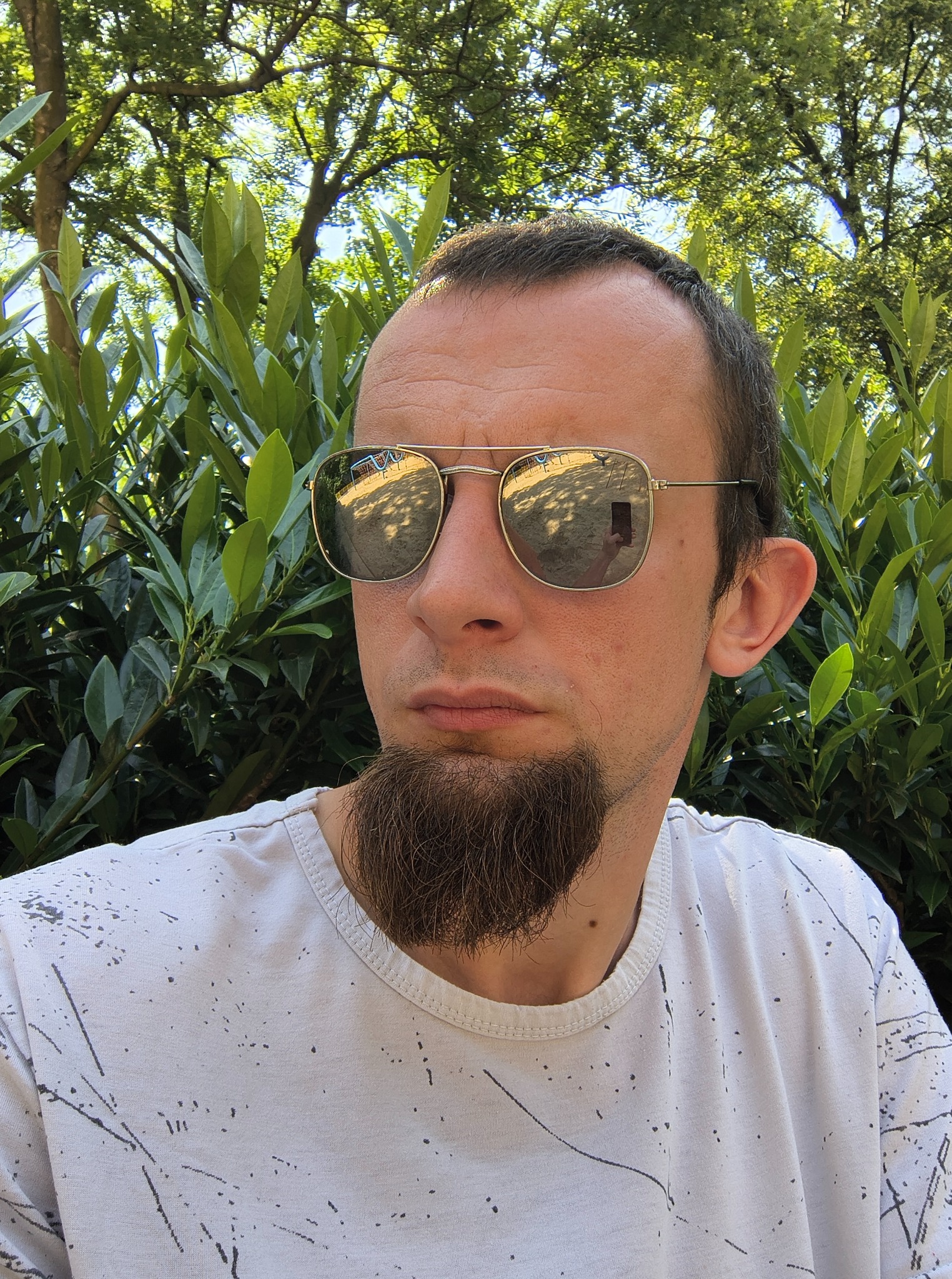 Mężczyzna w okularach przeciwsłonecznych, z nietypową brodą, na tle zielonych liści. W odbiciu okularów widoczny plac zabaw.