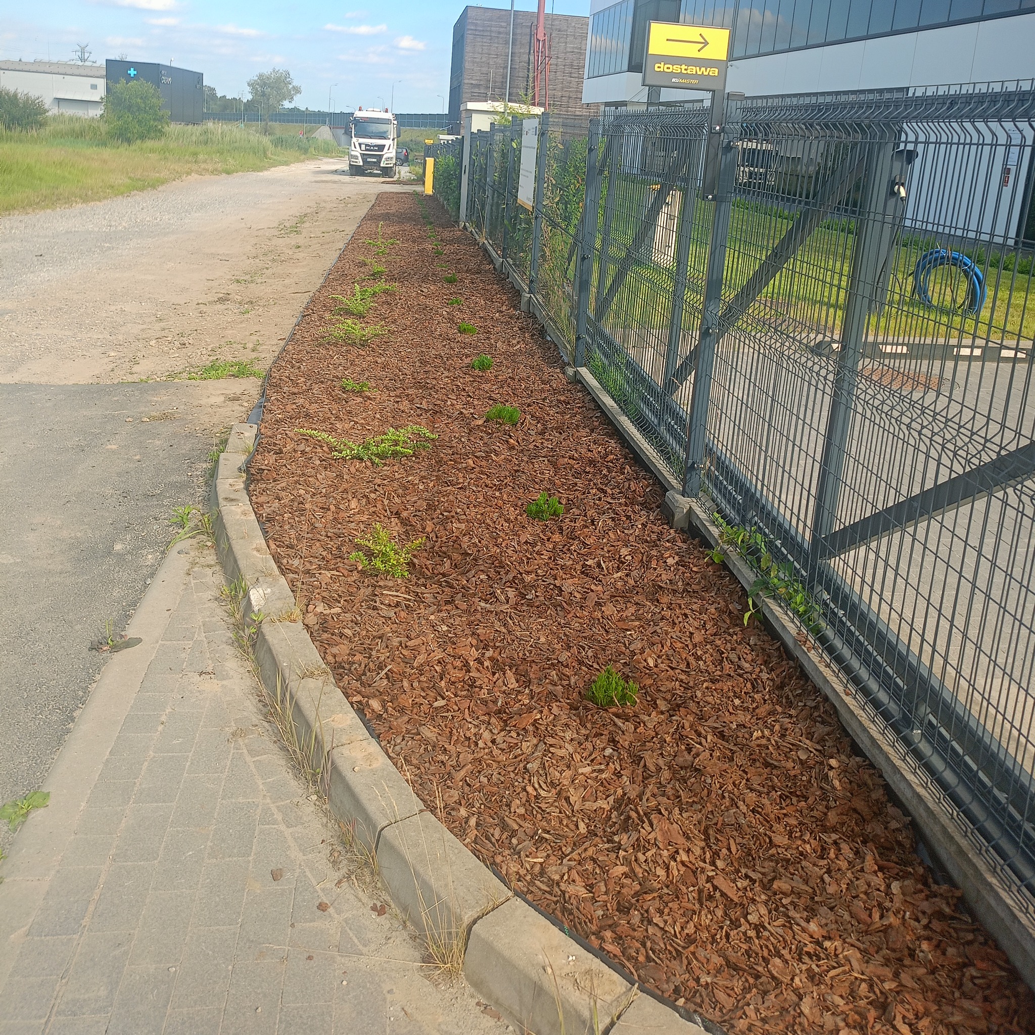 Rabata z kory i młodymi roślinami wzdłuż ogrodzenia panelowego, obok chodnika z kostki. W tle budynki przemysłowe i pojazd dostawczy.