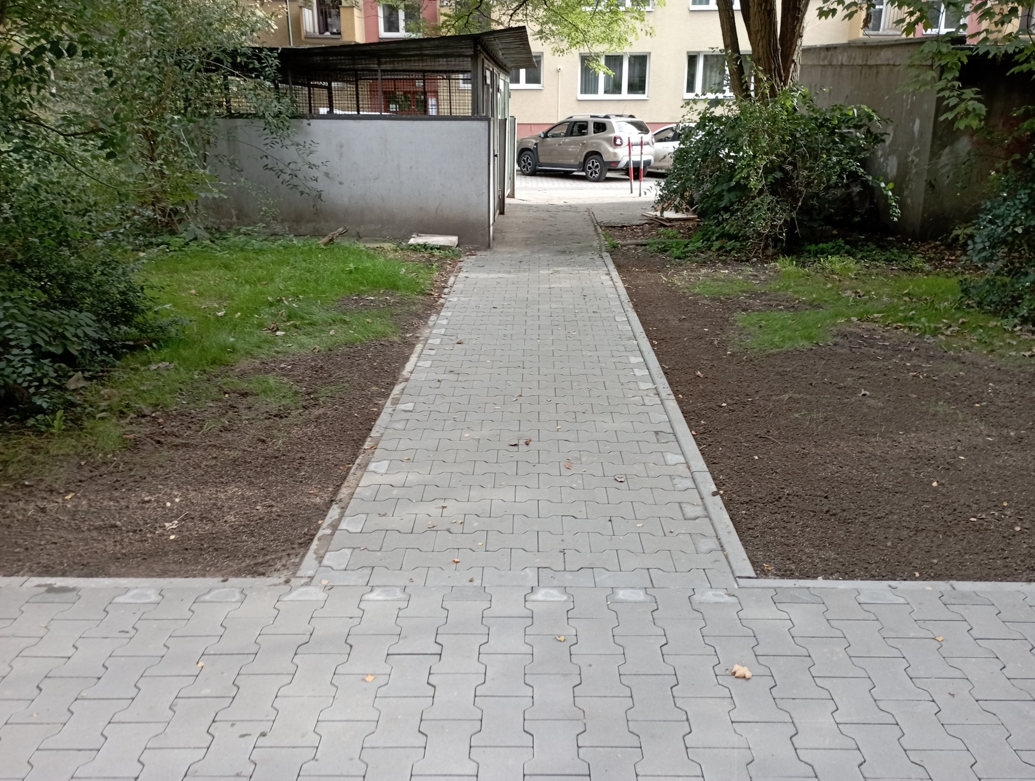 Ścieżka z kostki brukowej prowadząca przez trawnik z przygotowaną ziemią pod zasiew, obok budynek gospodarczy i zaparkowane auto w tle.