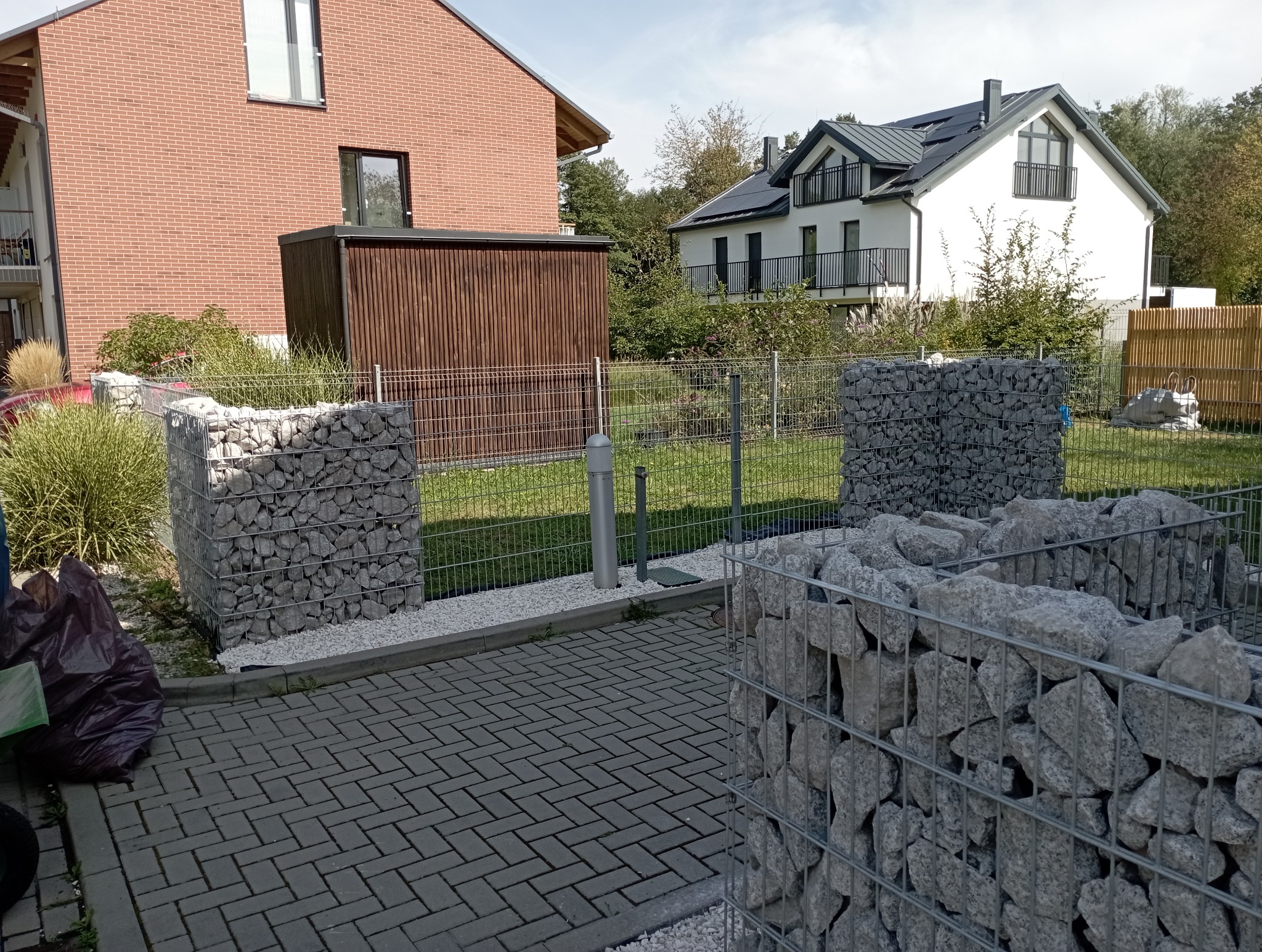 Gabiony z kamienia na posesji, tworzące element dekoracyjny ogrodu. W tle budynki mieszkalne i zieleń. Widoczna kostka brukowa i ogrodzenie.