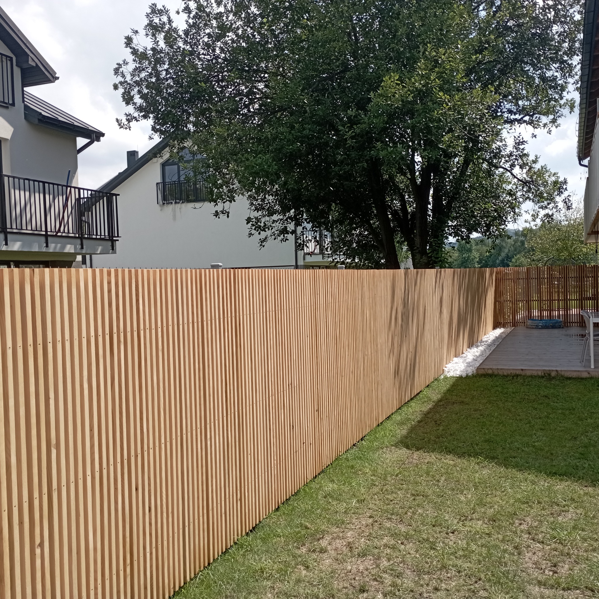 Nowoczesne ogrodzenie z pionowych desek w ogrodzie, z widoczną trawą i elementami architektury domu w tle. Jasne drewno ogrodzenia kontrastuje z zielenią trawnika.