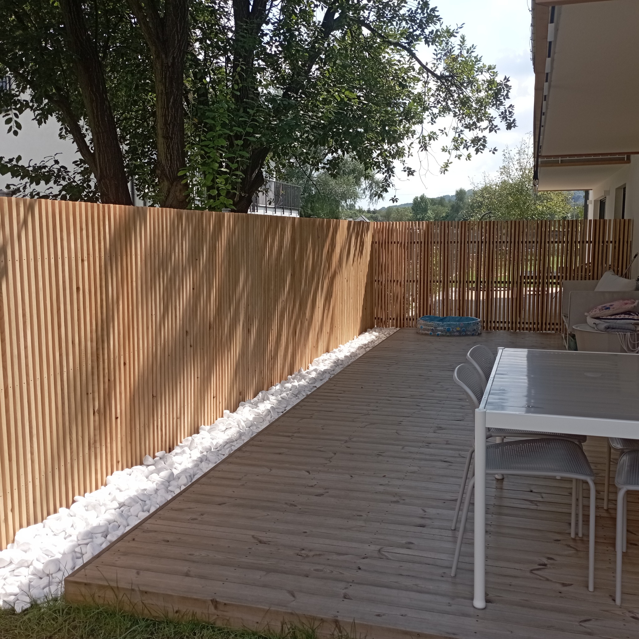 Drewniany taras z ażurowym ogrodzeniem i białymi kamieniami dekoracyjnymi. W tle zieleń drzew i krzewów, meble ogrodowe. Słoneczny dzień.