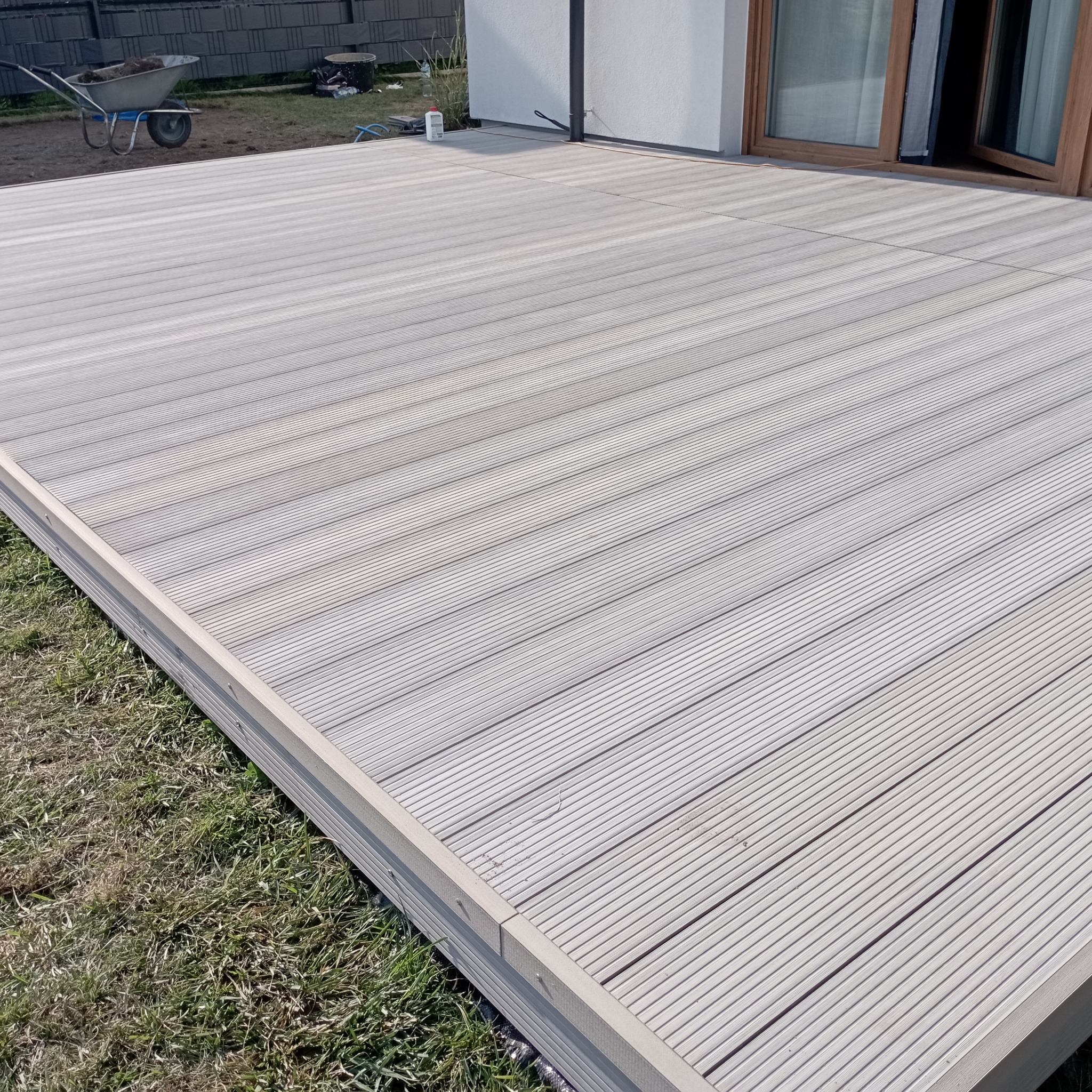 Jasnoszara deska tarasowa z kompozytu, ułożona przy nowoczesnym domu. W tle taczka z ziemią i częściowo widoczne okno.