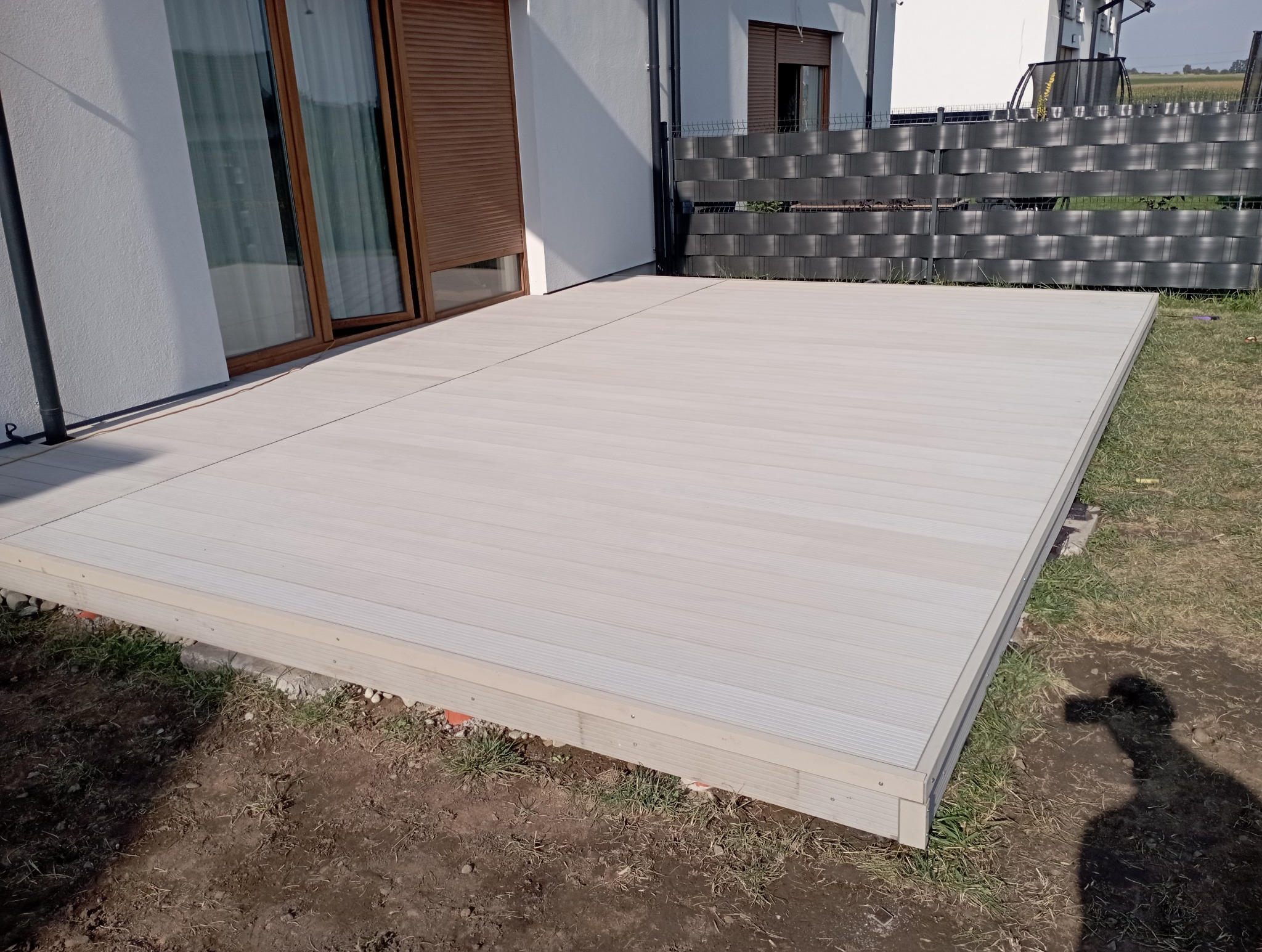 Nowoczesny taras z desek kompozytowych w odcieniach szarości, przylegający do białego budynku z brązowymi okiennicami. Widoczny fragment trawnika i ogrodzenia.