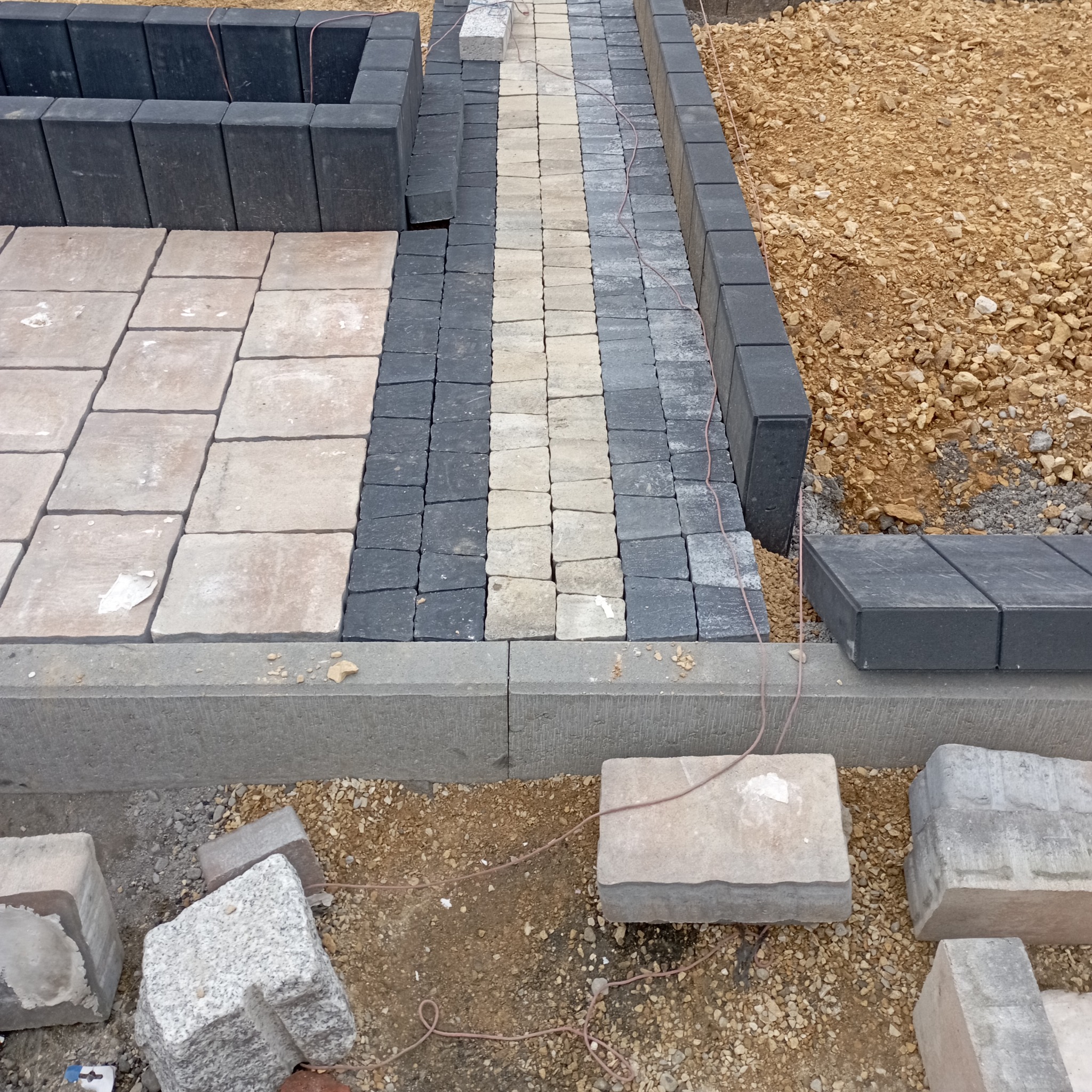Układanie kostki brukowej w ogrodzie, widok z góry. Fragment ścieżki z kostki w odcieniach szarości i beżu, obrzeża z ciemnej kostki, teren przygotowany pod dalsze prace.