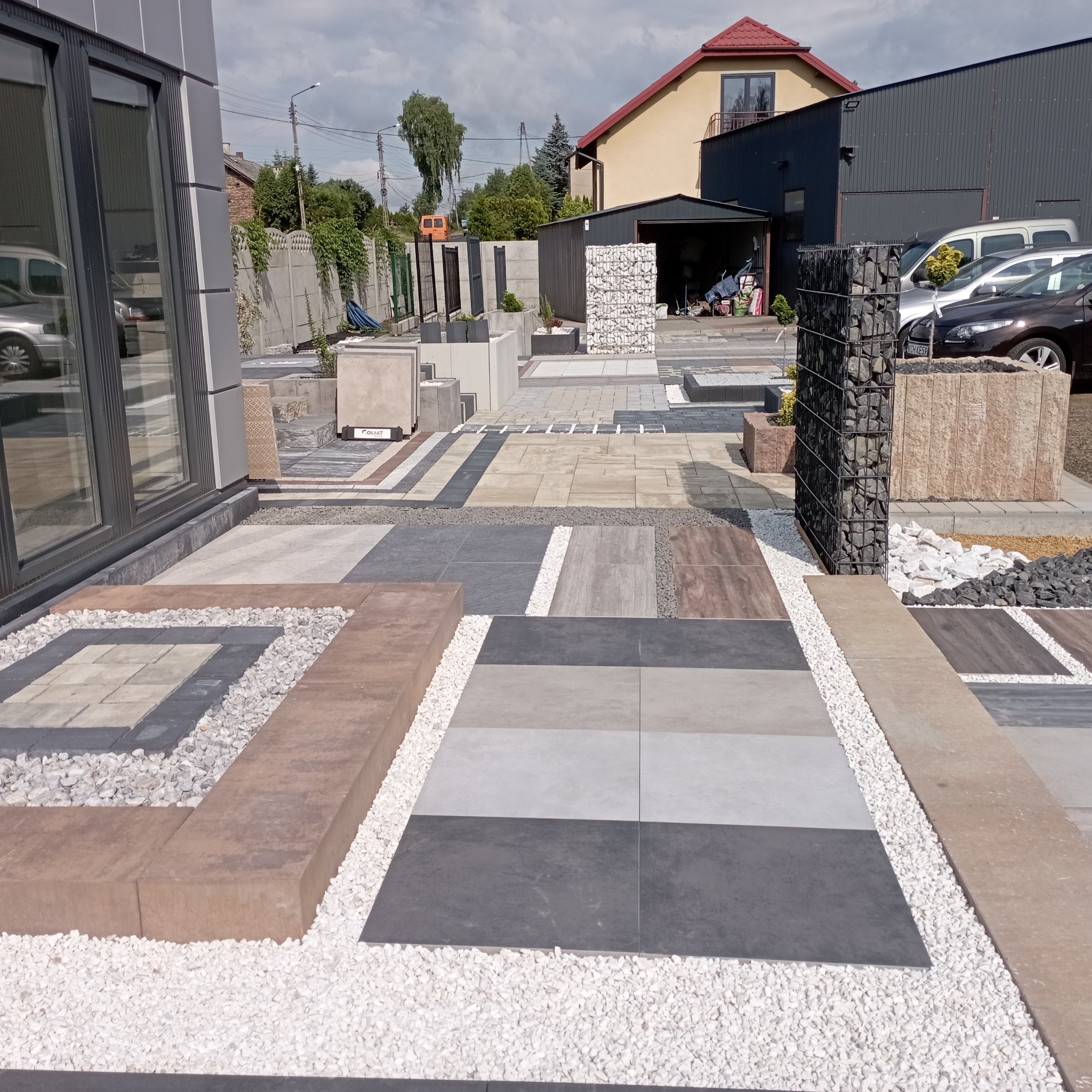 Ekspozycja różnorodnych wzorów kostki brukowej i płyt chodnikowych z białym grysem, prezentowana na zewnątrz budynku w słoneczny dzień. Widoczne elementy architektury.