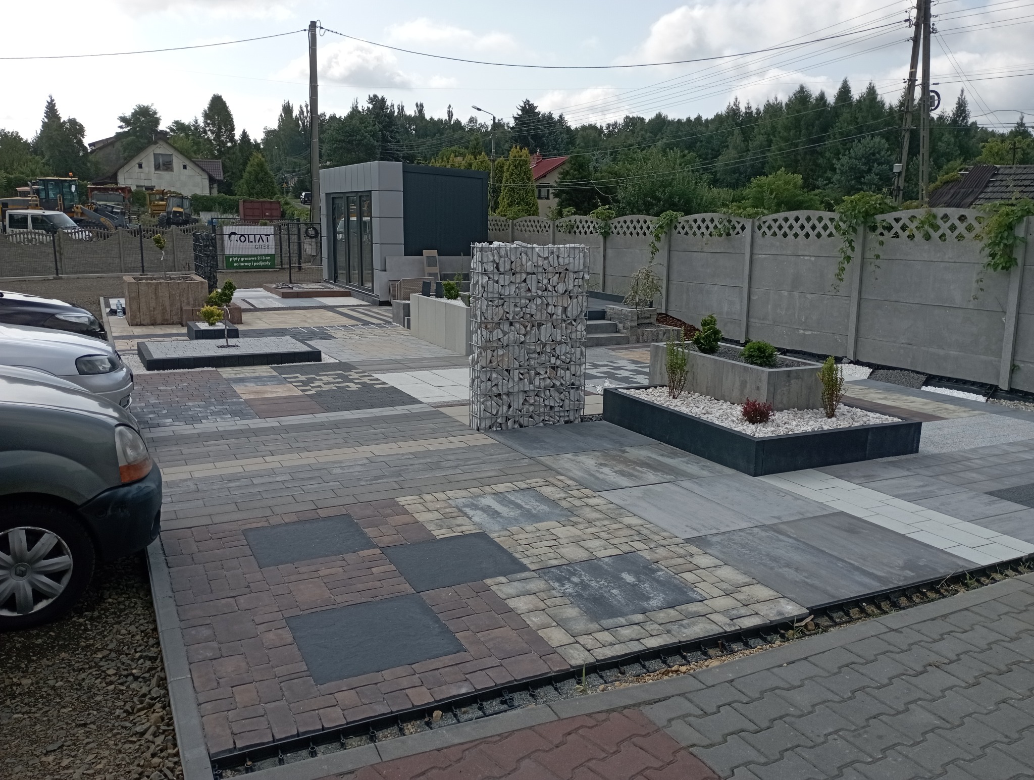 Ekspozycja kostki brukowej o różnych wzorach i kolorach, z elementami małej architektury ogrodowej, takimi jak gabiony i donice, na tle zieleni i zabudowań.