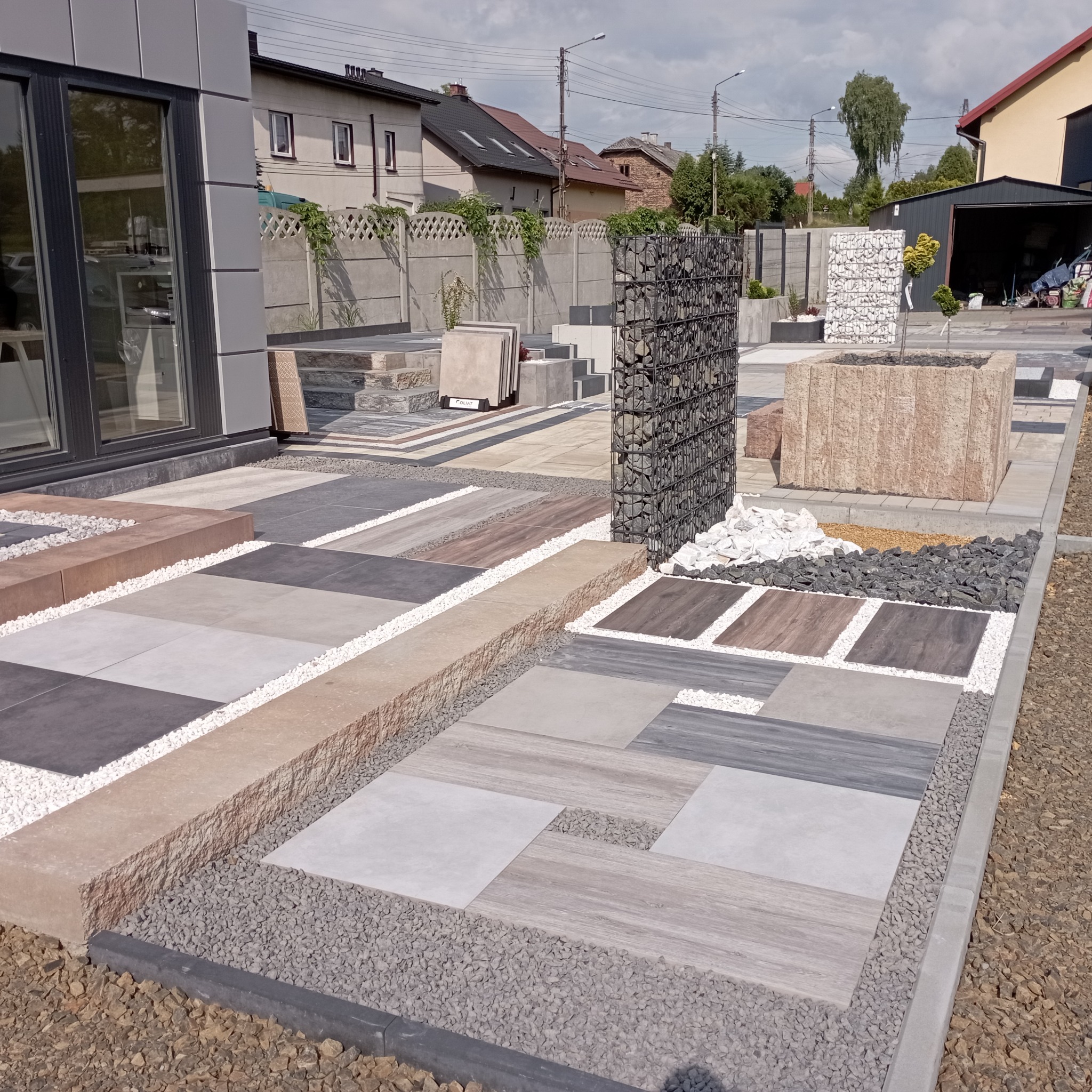 Ekspozycja różnorodnych wzorów i kolorów płyt chodnikowych, kostki brukowej i elementów dekoracyjnych, w tym gabionów, na terenie firmy.