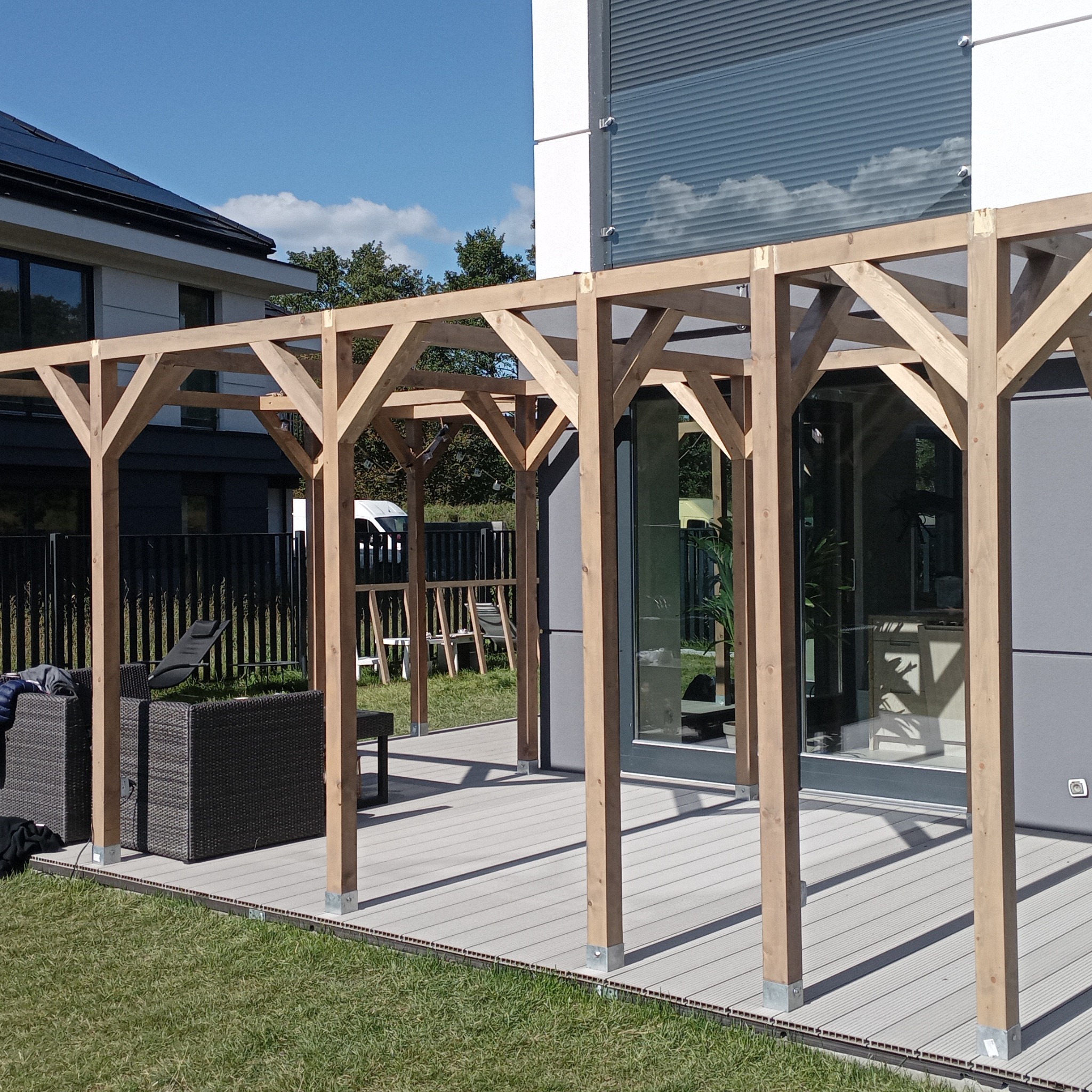 Drewniana pergola na tarasie z kompozytowych desek, przy nowoczesnym domu z dużymi oknami. Widoczne meble ogrodowe i trawnik. Słoneczny dzień.