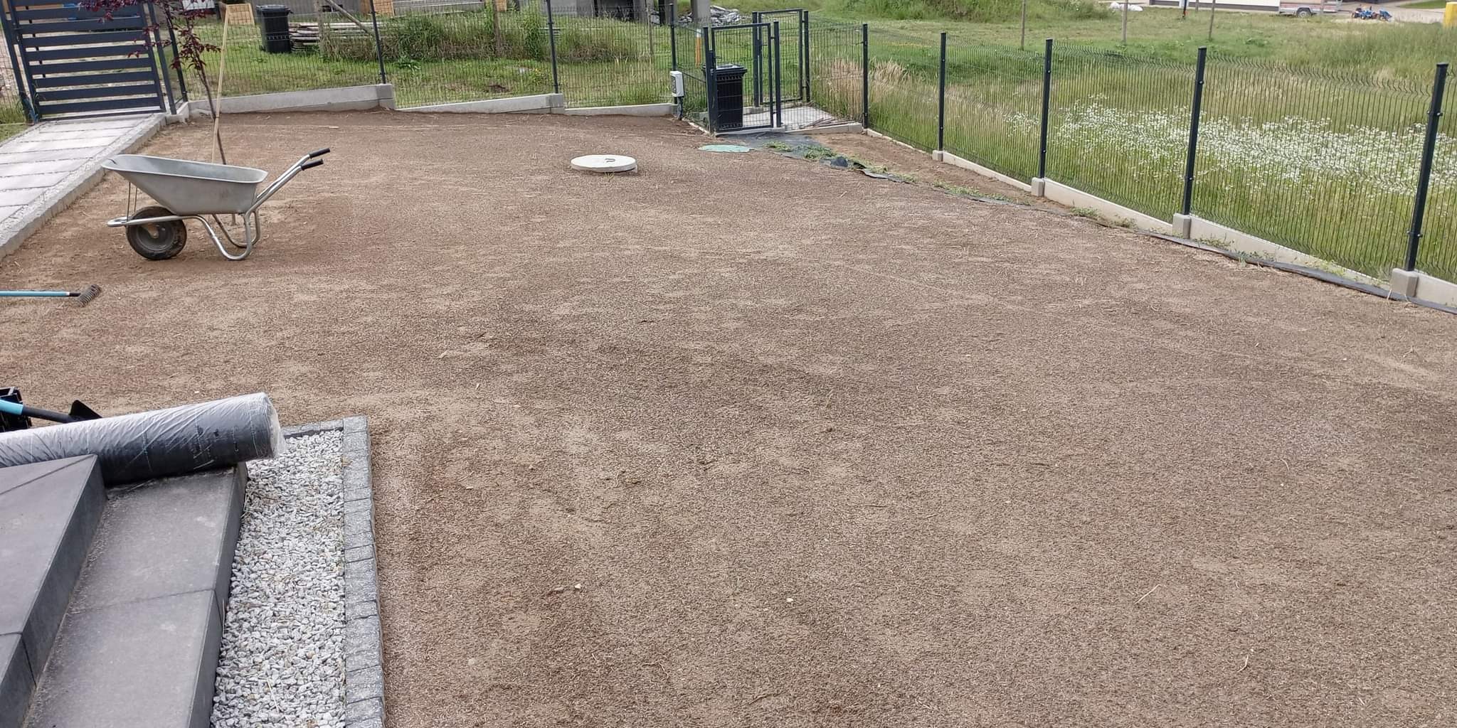 Przygotowany teren pod trawnik z widoczną taczką, zrolowaną trawą i nowym ogrodzeniem. W tle domy w zabudowie szeregowej.