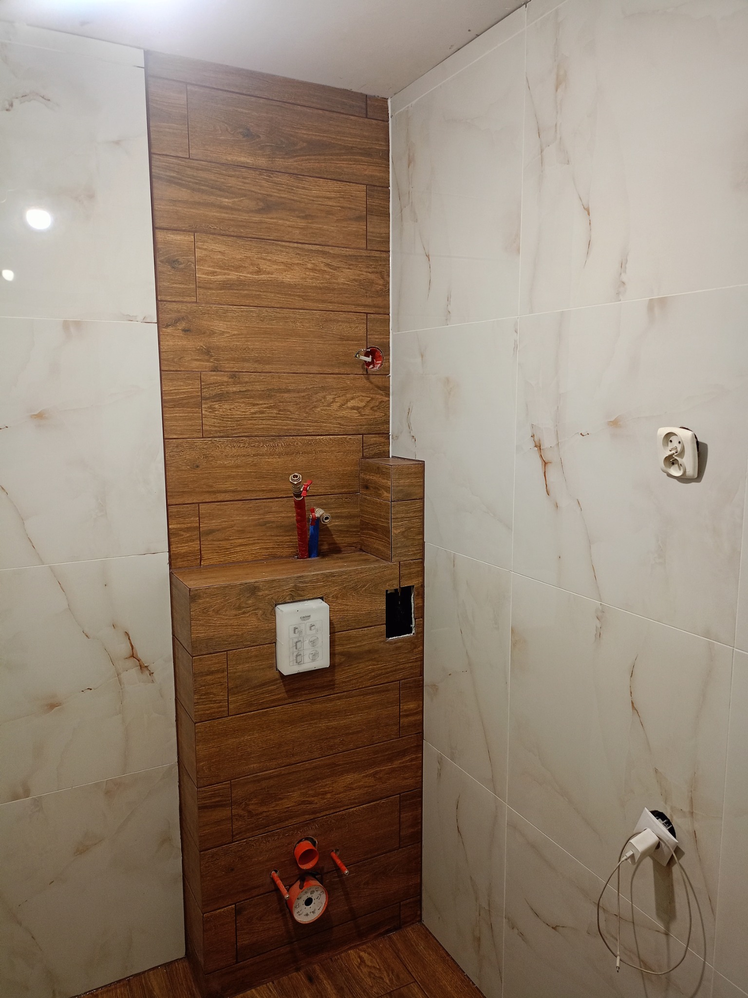 Remont łazienki: zabudowa WC z brązowych płytek imitujących drewno, białe marmurowe płytki na ścianach, widoczne instalacje wodne i elektryczne w trakcie prac.