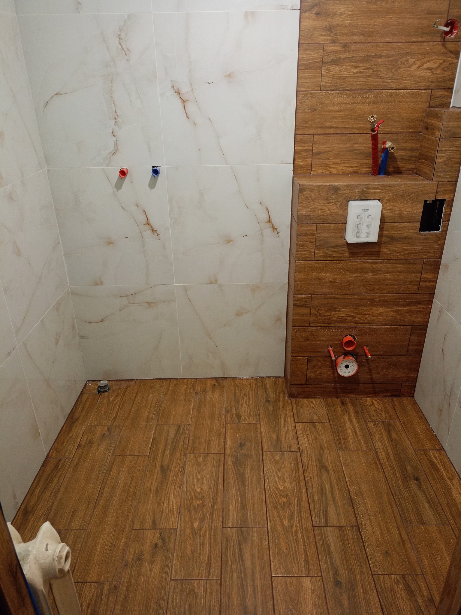 Wykończona łazienka z brązowymi płytkami imitującymi drewno na podłodze i ścianie, oraz białymi marmurkowymi płytkami na pozostałych ścianach. Widoczne instalacje wodne.