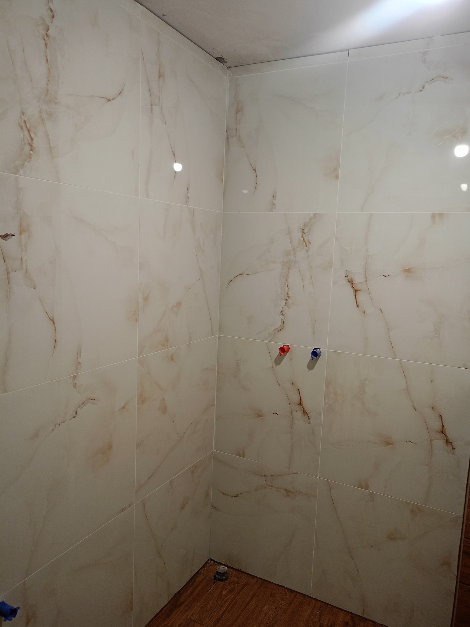 Wykończona łazienka z marmurowymi płytkami na ścianach i drewnopodobną podłogą. Widoczne przyłącza wodne w kolorze czerwonym i niebieskim. Minimalistyczny design.