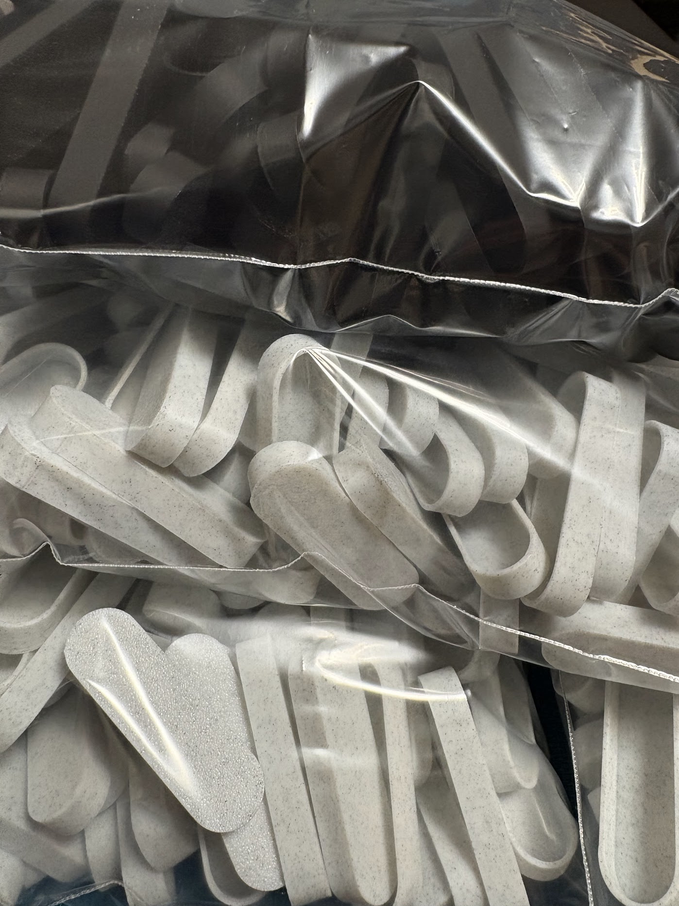 Szare, plastikowe elementy w przezroczystych workach foliowych. Elementy o nieregularnym kształcie, przypominające uchwyty, z widoczną fakturą materiału.