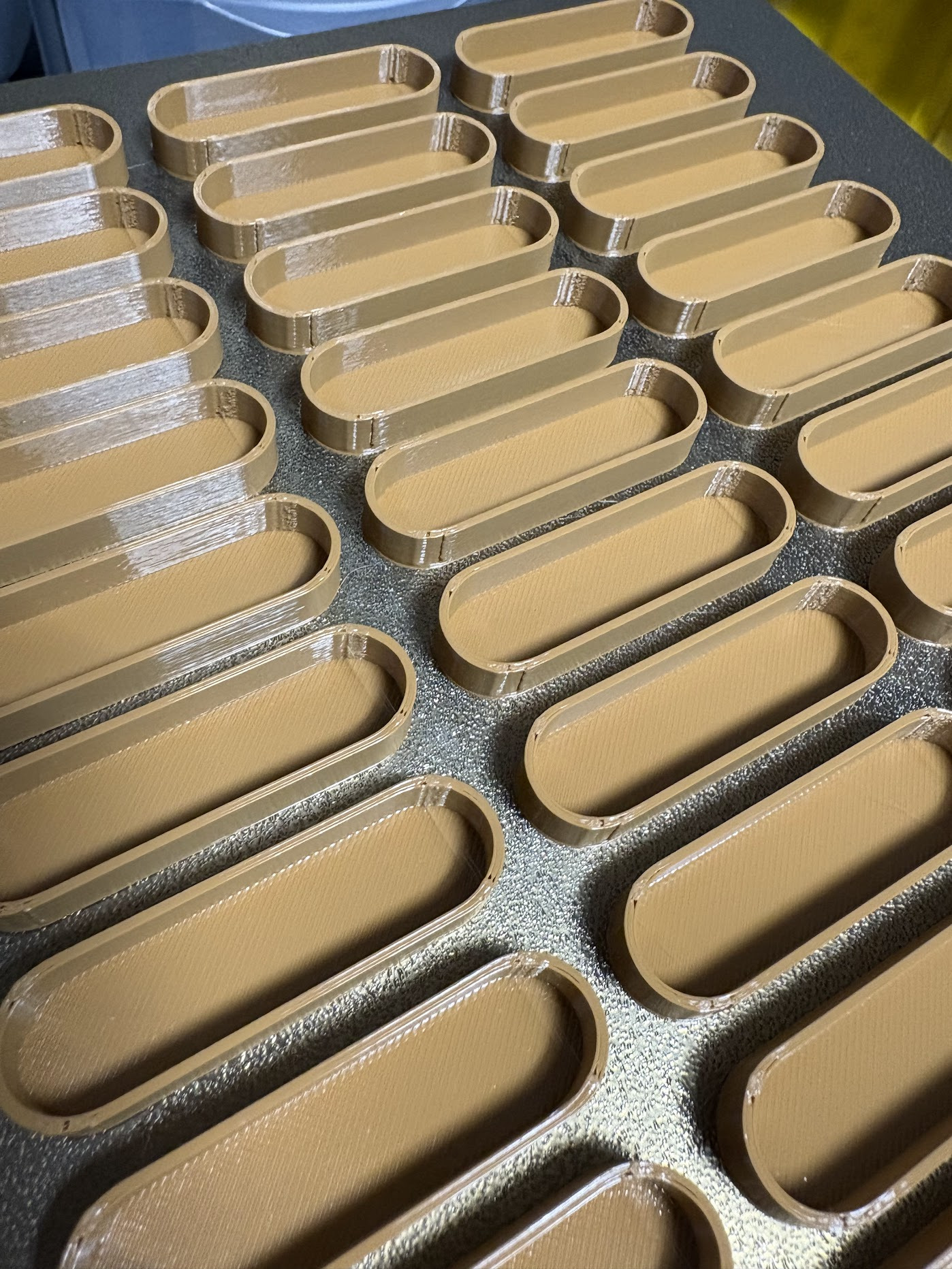 Seria beżowych, owalnych pojemników z tworzywa sztucznego, ułożonych równolegle na szarej powierzchni. Widoczna tekstura warstw, sugerująca proces wytwarzania.