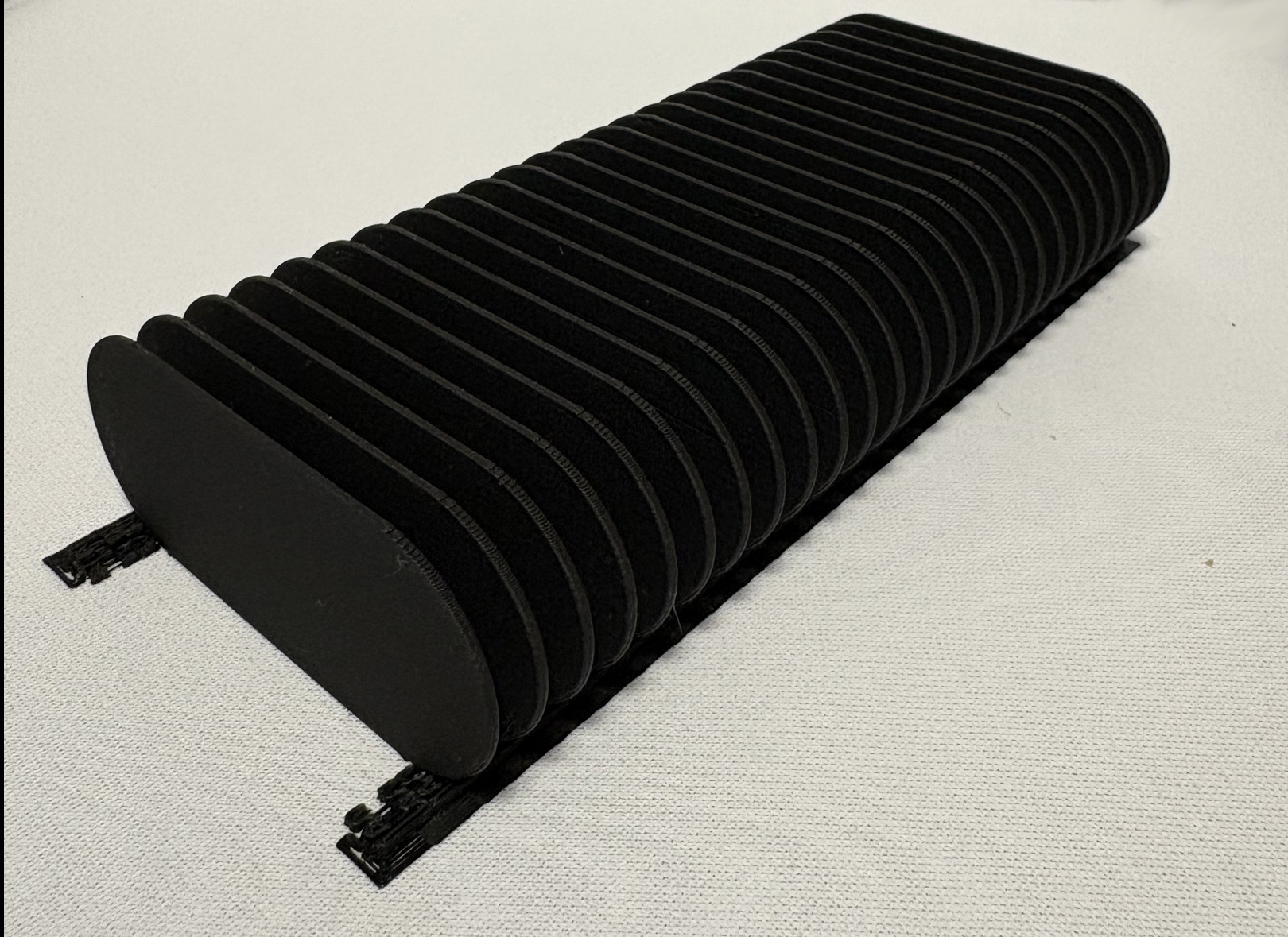 Czarny, żebrowany element z tworzywa sztucznego, prawdopodobnie osłona, z widocznymi śladami warstw, leżący na białej powierzchni. Detal wykonany z precyzją.