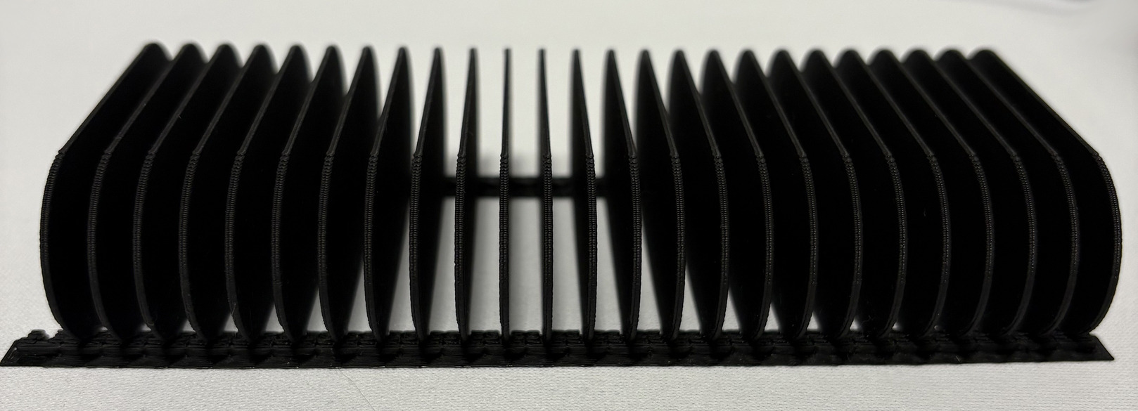 Czarny, żebrowany element konstrukcyjny z tworzywa sztucznego, widoczny z boku na białym tle. Widoczne ślady warstwowego wykonania.