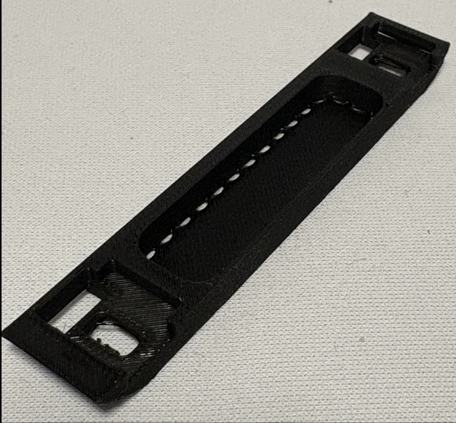 Czarny, podłużny element z tworzywa sztucznego z otworami i wgłębieniami, prawdopodobnie wykonany metodą druku 3D, leżący na szarej powierzchni.
