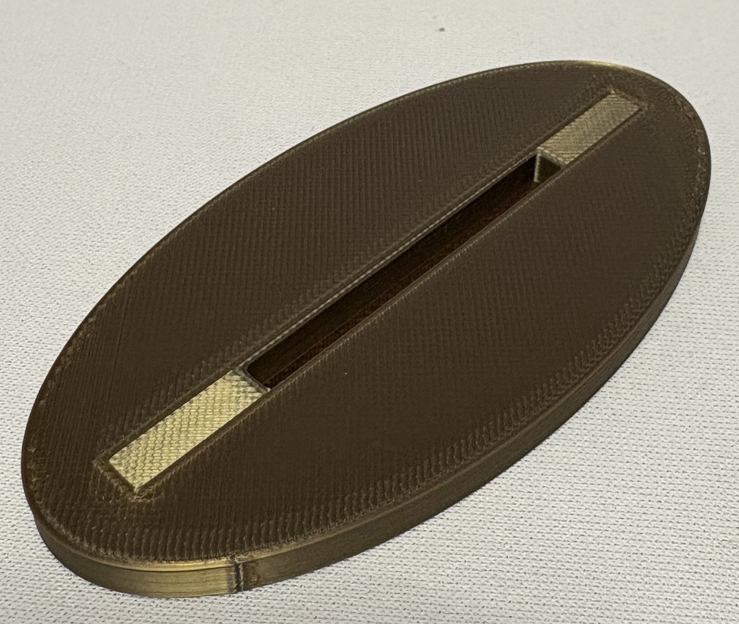 Brązowa, owalna tsuba (jelec) do japońskiego miecza, z prostokątnym otworem i metalowymi wstawkami, leżąca na jasnym tle.