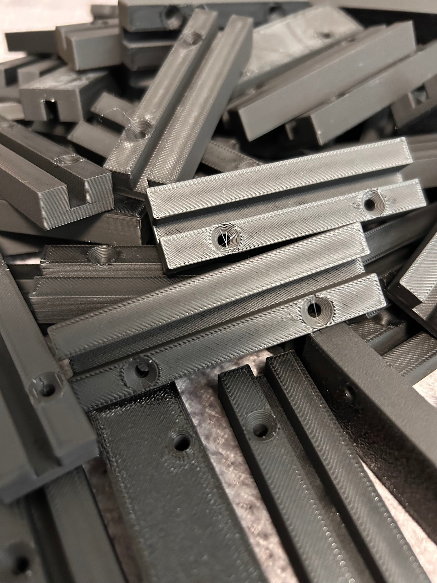 Szare, prostokątne elementy z otworami ułożone warstwami, widoczne ślady obróbki, co sugeruje wykonanie metodą addytywną.