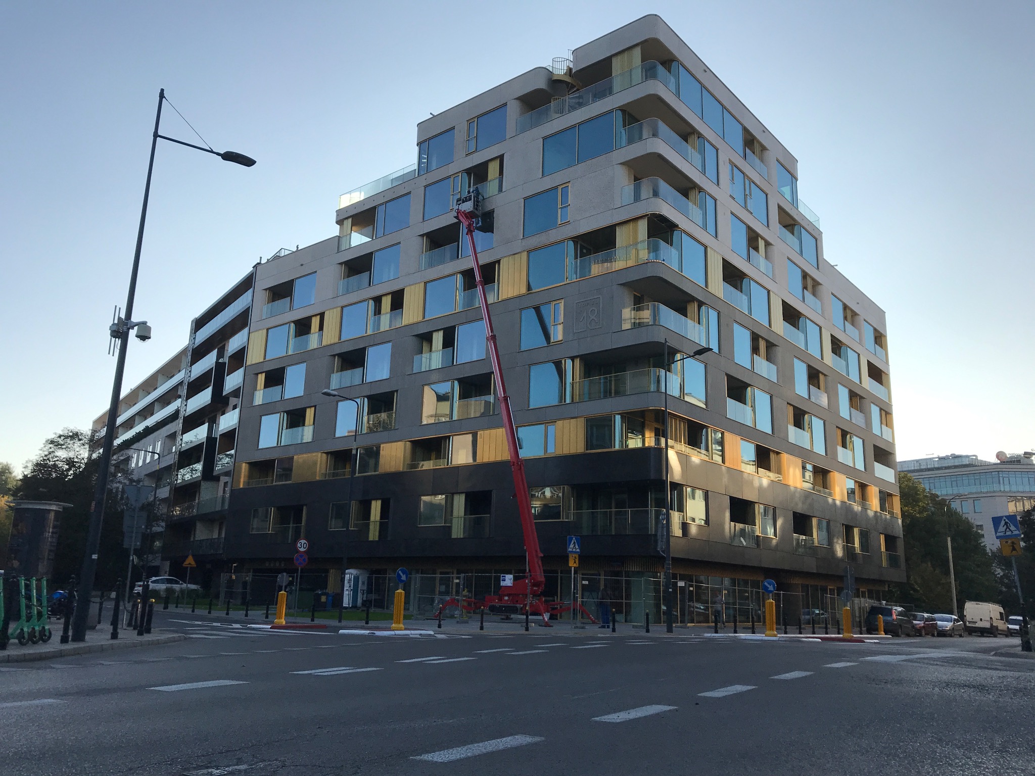 Nowoczesny budynek wielorodzinny z podnośnikiem koszowym przy elewacji. Widoczne złote elementy wykończenia i duże okna. Ulica z oznakowaniem w tle.