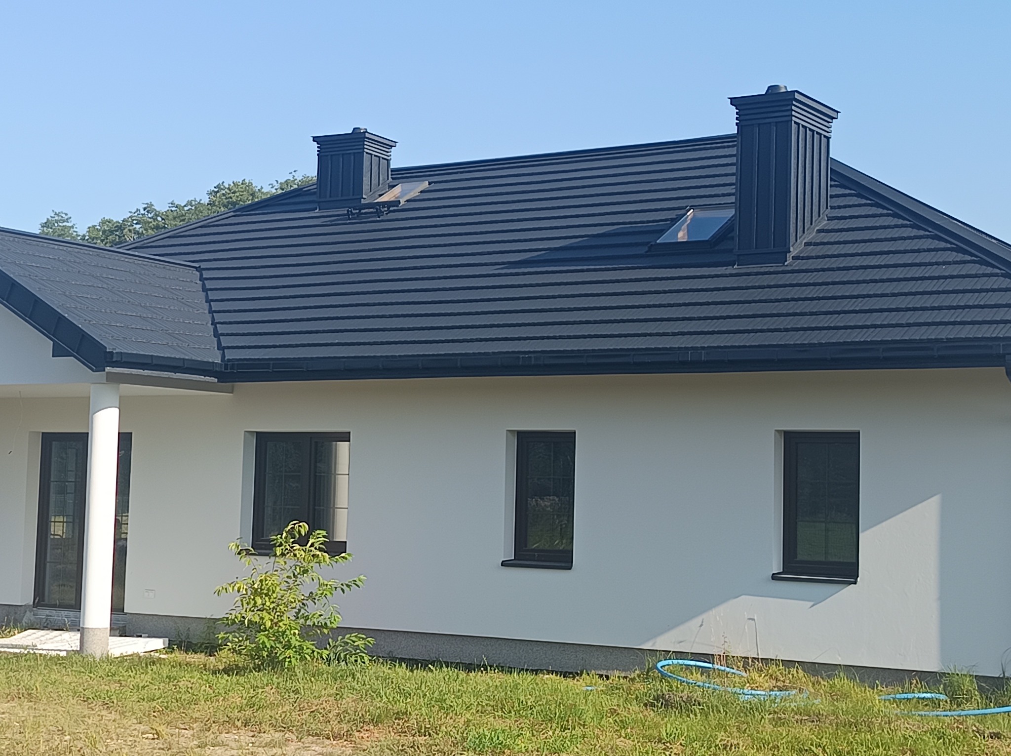 Czarny dach z dachówki modułowej na białym domu jednorodzinnym z czarnymi oknami, widoczne kominy i okno dachowe, w tle drzewa i błękitne niebo.