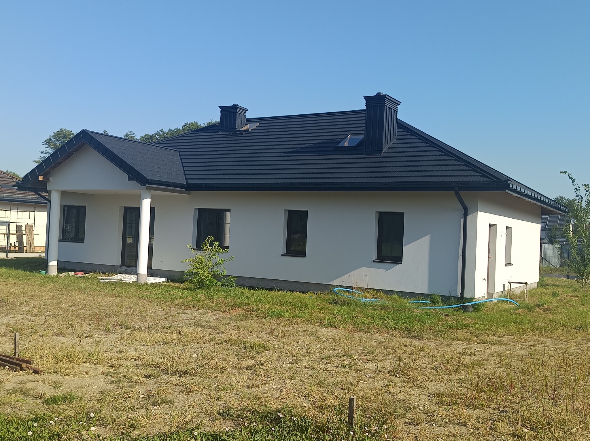 Nowoczesny dom jednorodzinny z ciemną dachówką i dwoma kominami. Biała elewacja, widok z boku, trawnik z fragmentami piasku.