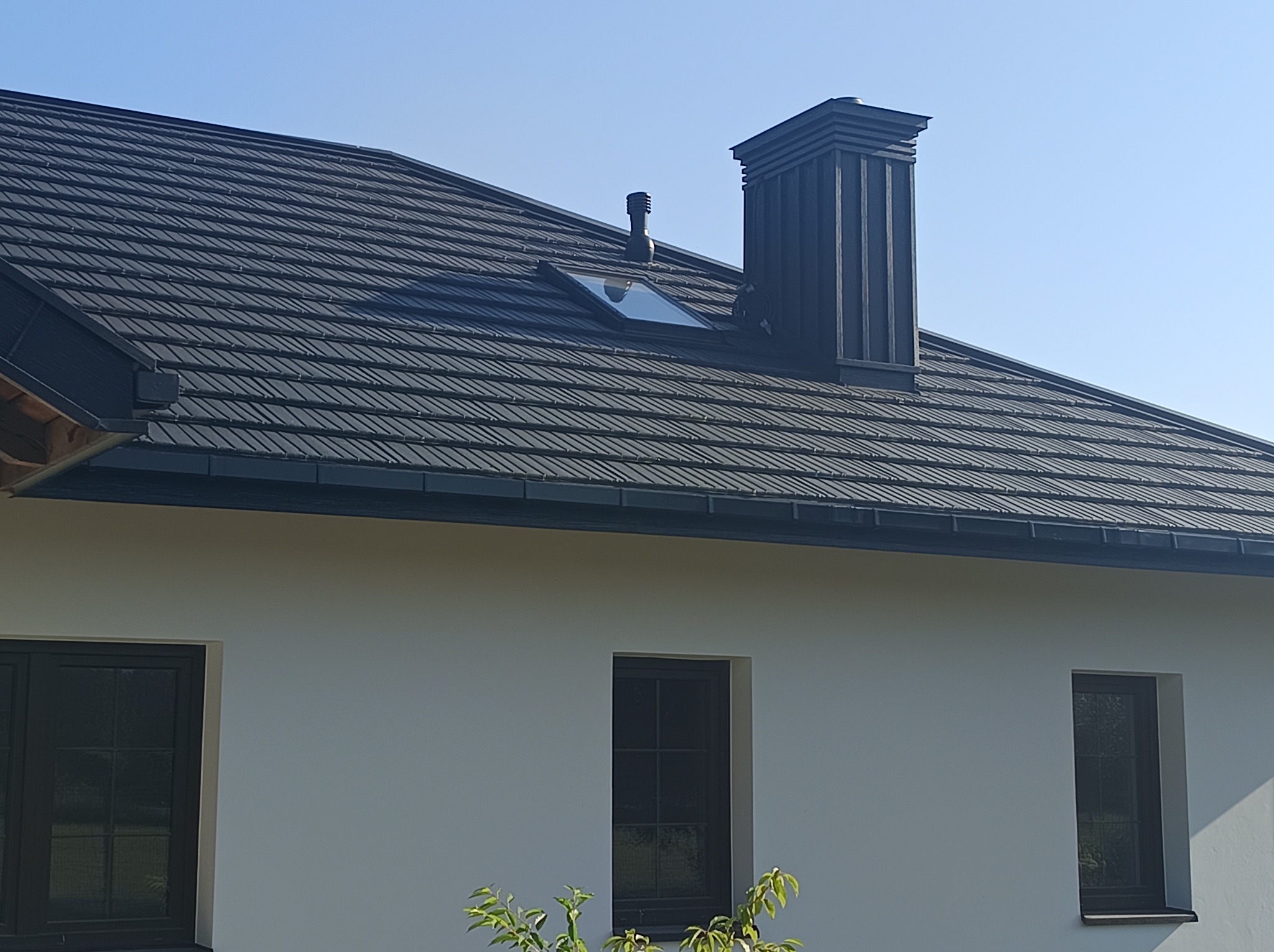 Czarny dach z blachy trapezowej, komin z obróbką blacharską i okno dachowe na tle błękitnego nieba, fragment ściany budynku z oknami.