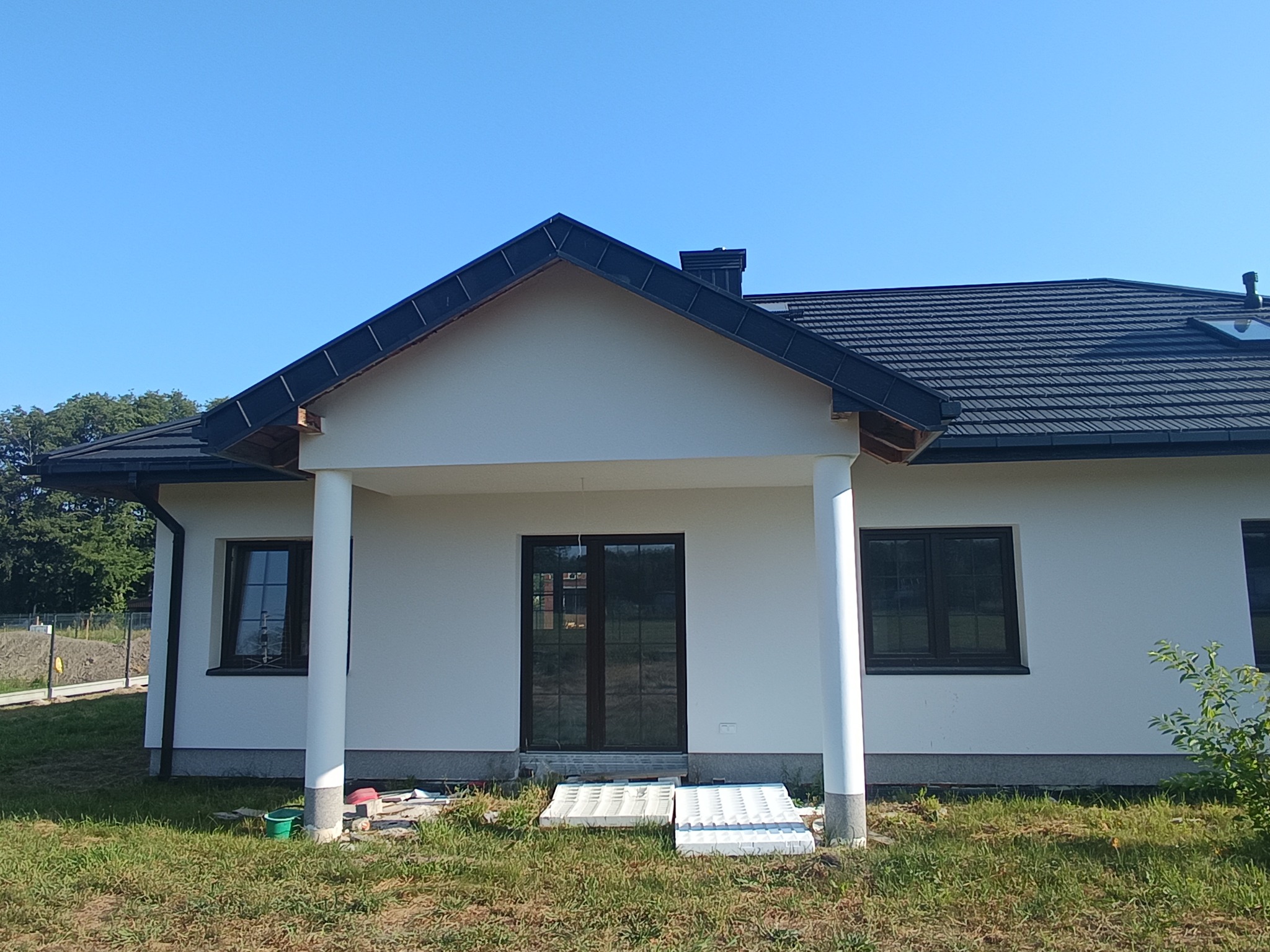 Dom jednorodzinny z ciemnym dachem z blachy trapezowej i białymi ścianami. Widoczne okna, kolumny wejściowe i fragment ogrodu.