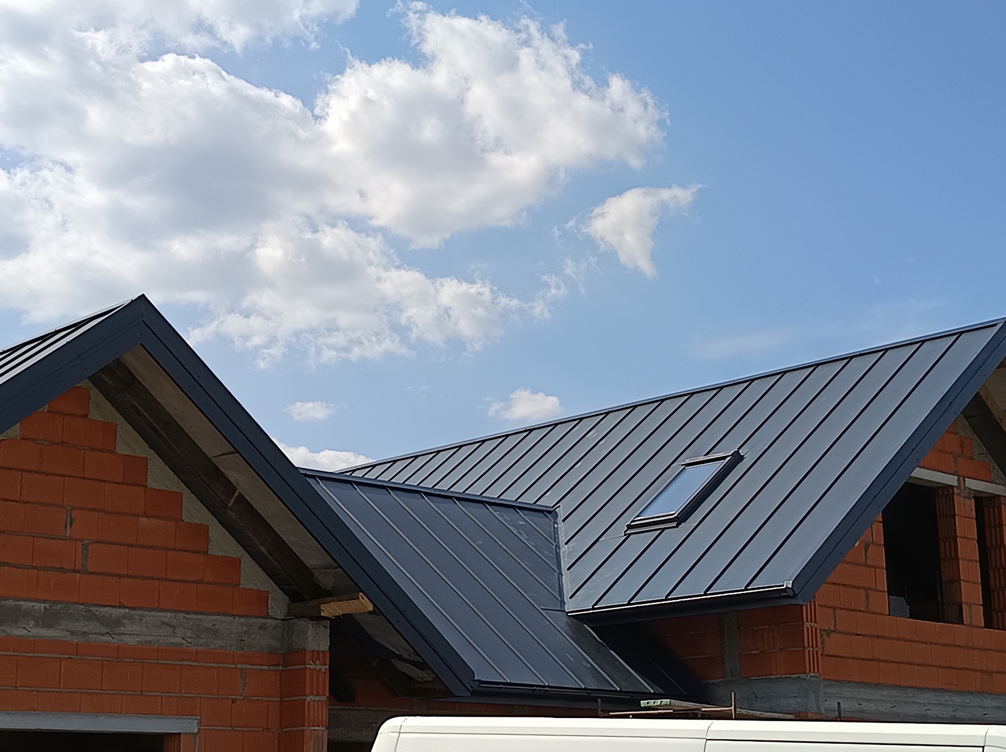 Nowoczesny dach z blachy na domu w trakcie budowy, widoczne okno dachowe i fragment nieotynkowanej ściany z czerwonej cegły. Błękitne niebo z chmurami w tle.