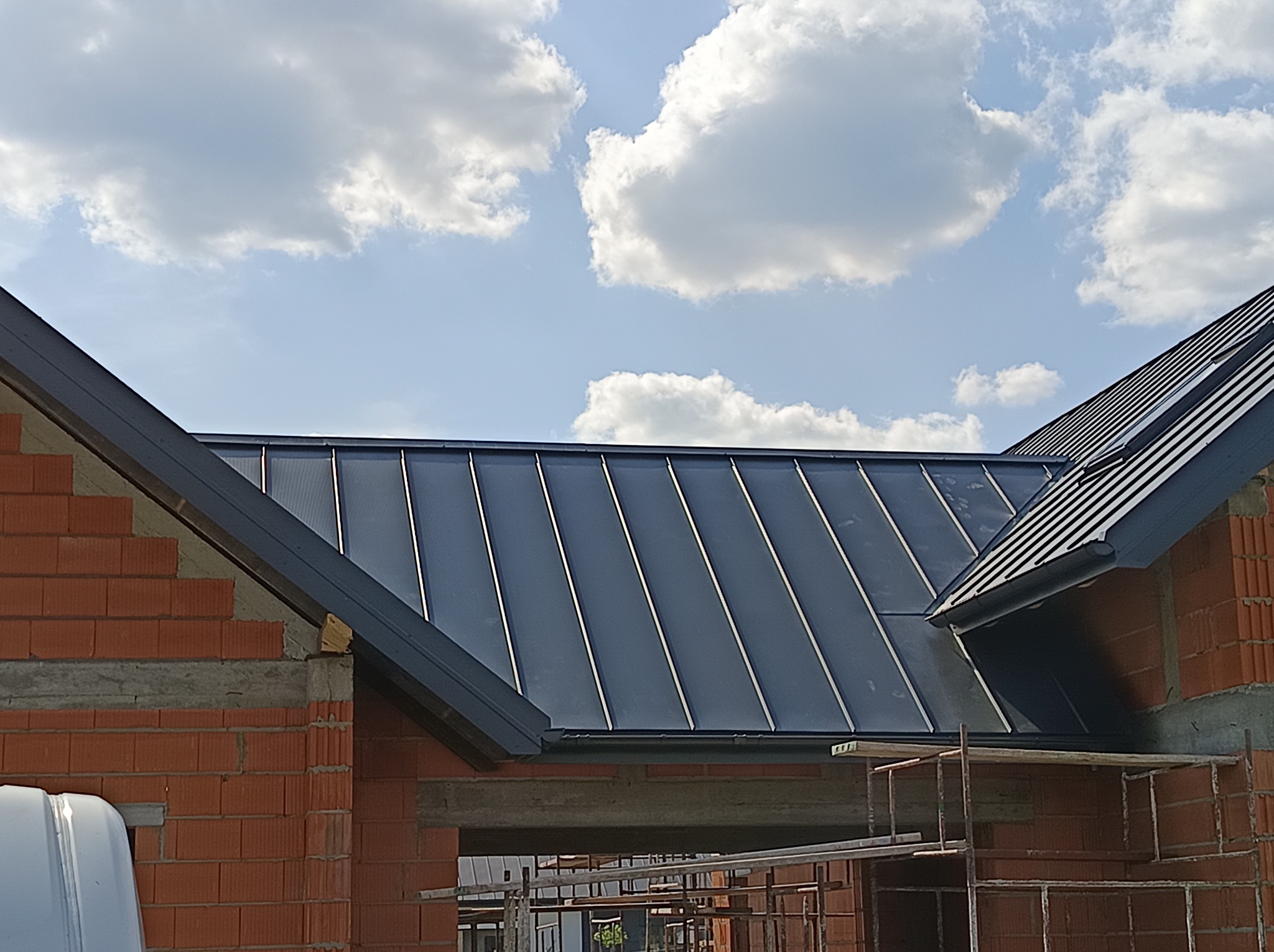 Krycie dachu blachą na rąbek stojący w Grodzisku Mazowieckim. Widoczne fragmenty ścian budynku w trakcie budowy i błękitne niebo z chmurami.