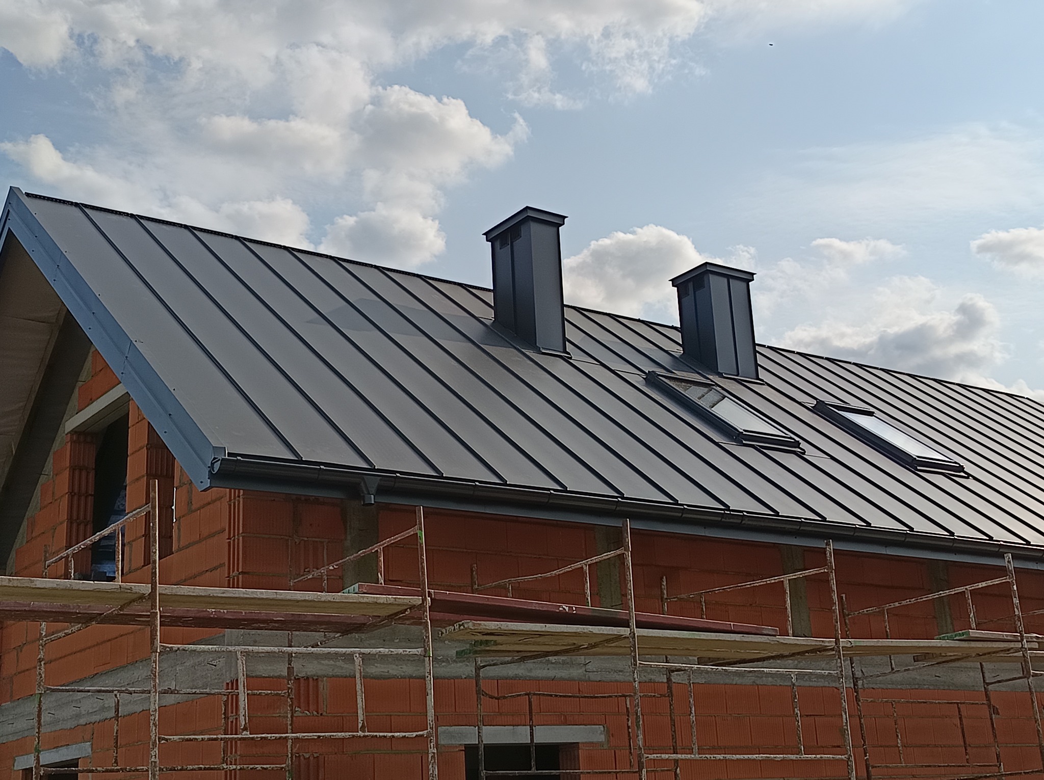 Nowy, grafitowy dach z blachy na budynku w trakcie budowy, widoczne okna dachowe i kominy. Ujęcie z dołu, na pierwszym planie rusztowanie.