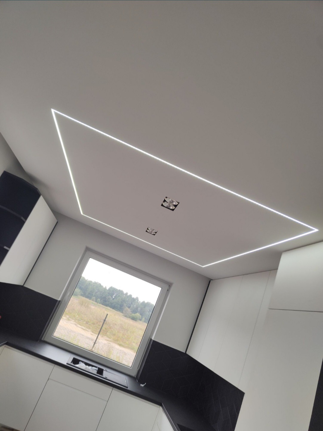 Nowoczesne oświetlenie LED w kształcie trapezu na suficie w kuchni. Widoczne okno z widokiem na zewnątrz. Minimalistyczny design wnętrza.