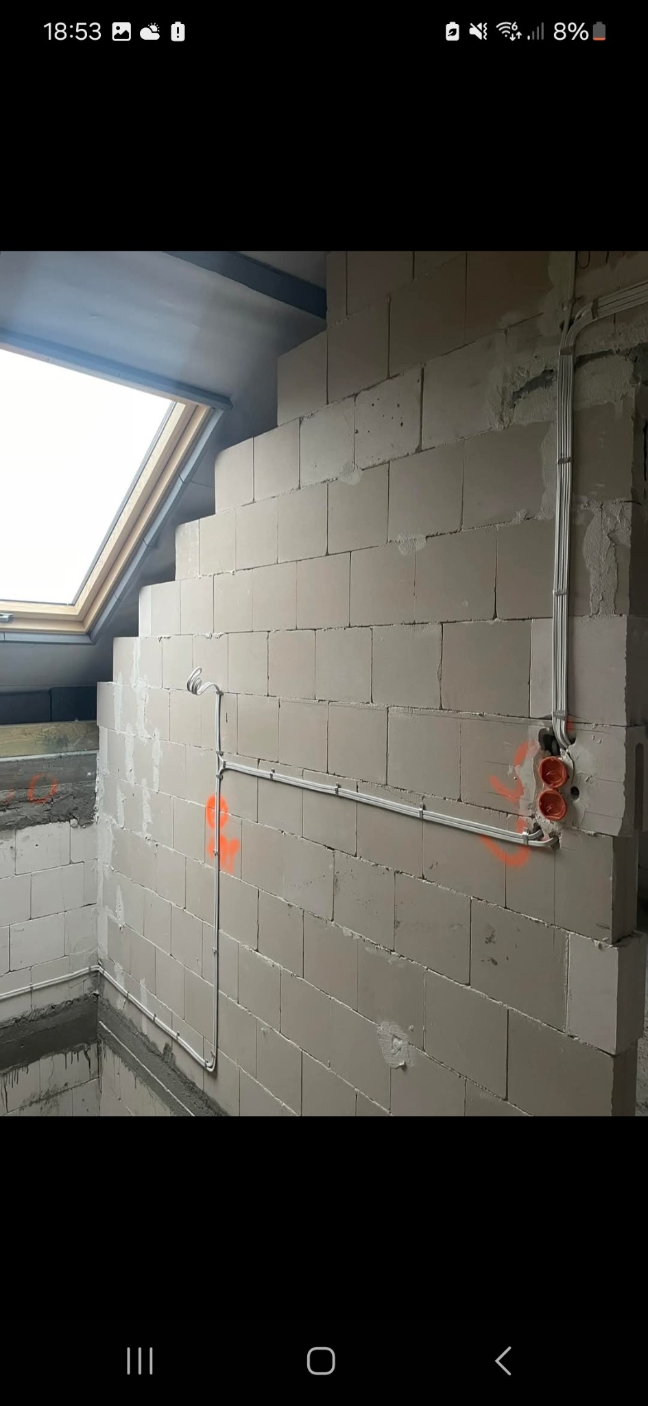 Surowa ściana z betonu komórkowego w trakcie budowy poddasza, z widoczną instalacją elektryczną w białych rurkach i puszkami. Okno dachowe w tle.