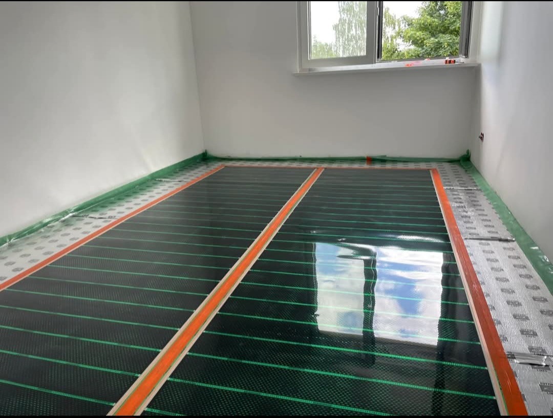 Montaż folii grzewczej na podłodze w pokoju z białymi ścianami i oknem. Widoczne zielone linie grzewcze i pomarańczowe paski mocujące. Prace instalacyjne w toku.