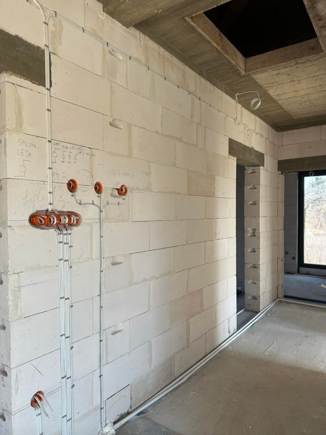 Surowy stan budynku z widoczną, częściowo rozprowadzoną instalacją elektryczną w białych rurkach na ścianie z betonu komórkowego. Widoczne punkty oświetleniowe.