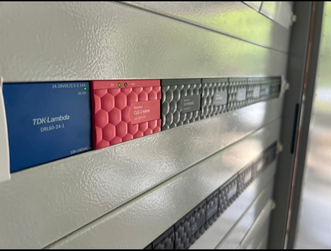 Szara szafa sterownicza z modułami: niebieski TDK-Lambda DRL60-24-1, czerwony Grenton CLU Z-Wave i szare moduły z etykietami. Widoczne diody sygnalizacyjne.