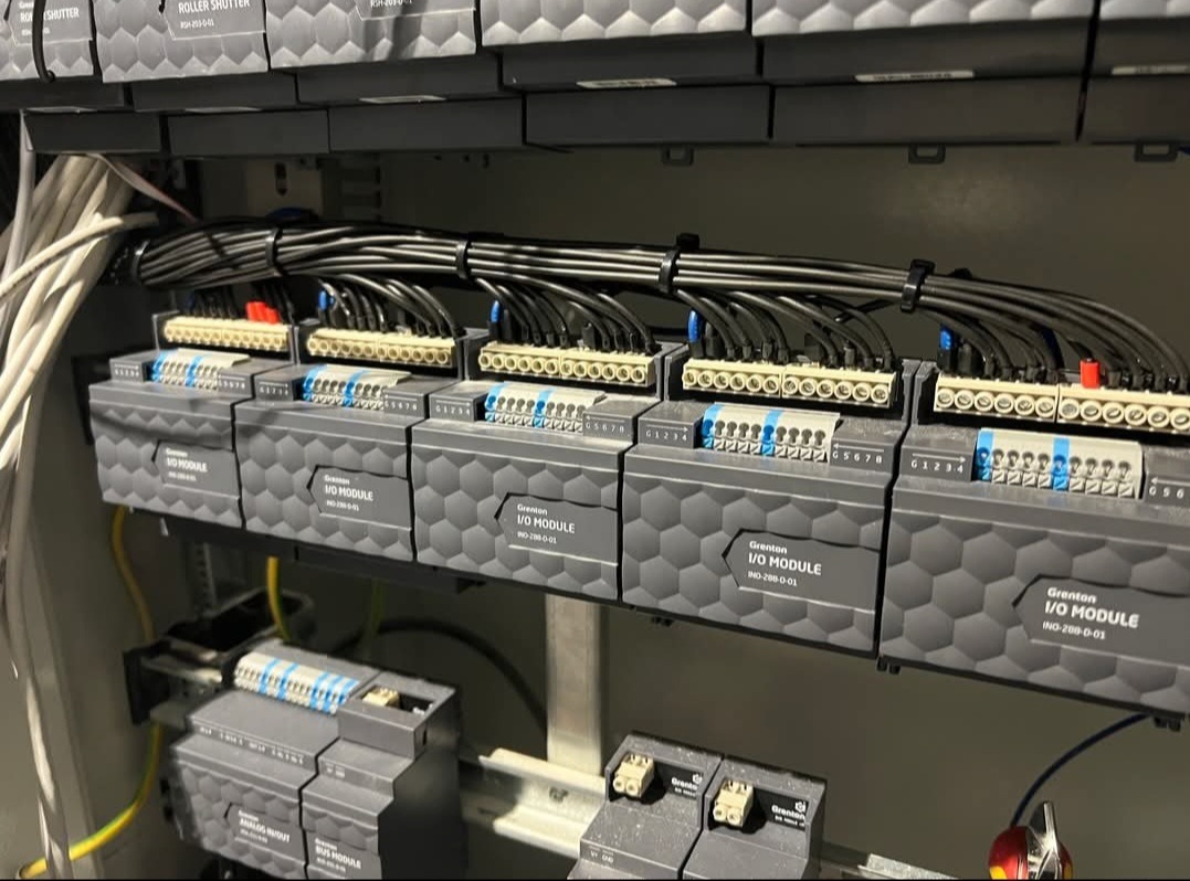 Szare moduły I/O Grenton INO-288-0-01 w szafie sterowniczej, z podłączonymi kablami, bliski kadr na instalację elektryczną.