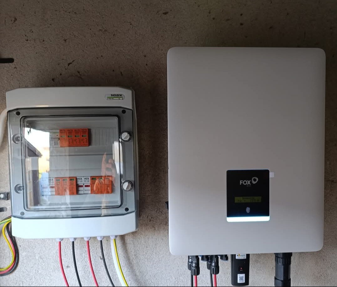 Zamontowana skrzynka z bezpiecznikami i falownik FOX ESS na ścianie. Widoczne przewody elektryczne. Detal instalacji fotowoltaicznej wewnątrz budynku.