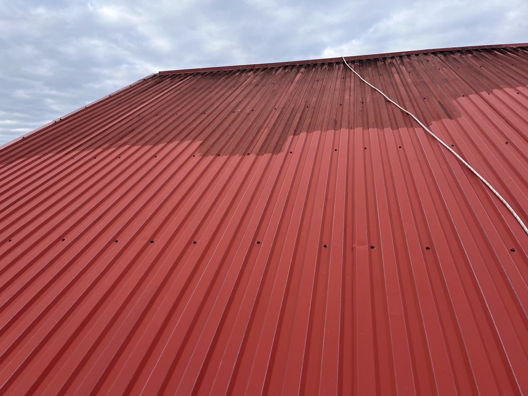 Częściowo umyty, czerwony dach z blachy trapezowej pod pochmurnym niebem. Widoczna lina. Porównanie czystej i zabrudzonej powierzchni.