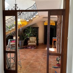 Ontera Design & Build - Montaż drewnianych drzwi przesuwnych w domu z terakotową podłogą. Widoczne narzędzia i schody z kutą balustradą. Praca w toku.