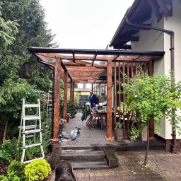 Ontera Design & Build - Drewniana pergola z zadaszeniem z poliwęglanu, przylegająca do domu, otoczona zielenią. Widoczne narzędzia i częściowo sylwetka osoby w tle, sugerujące etap budowy.
