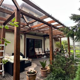 Ontera Design & Build - Drewniana pergola z przezroczystym zadaszeniem, meble ogrodowe na tarasie, drabina i roślinność. Widok na dom z zewnątrz, relaksacyjna przestrzeń na świeżym powietrzu.