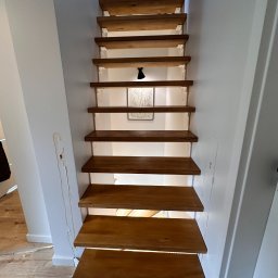 Ontera Design & Build - Wąskie schody drewniane w jasnym wnętrzu, widok od dołu. Stopnie oświetlone od spodu, na ścianie grafika w ramce. Nierówności na ścianie po lewej stronie.